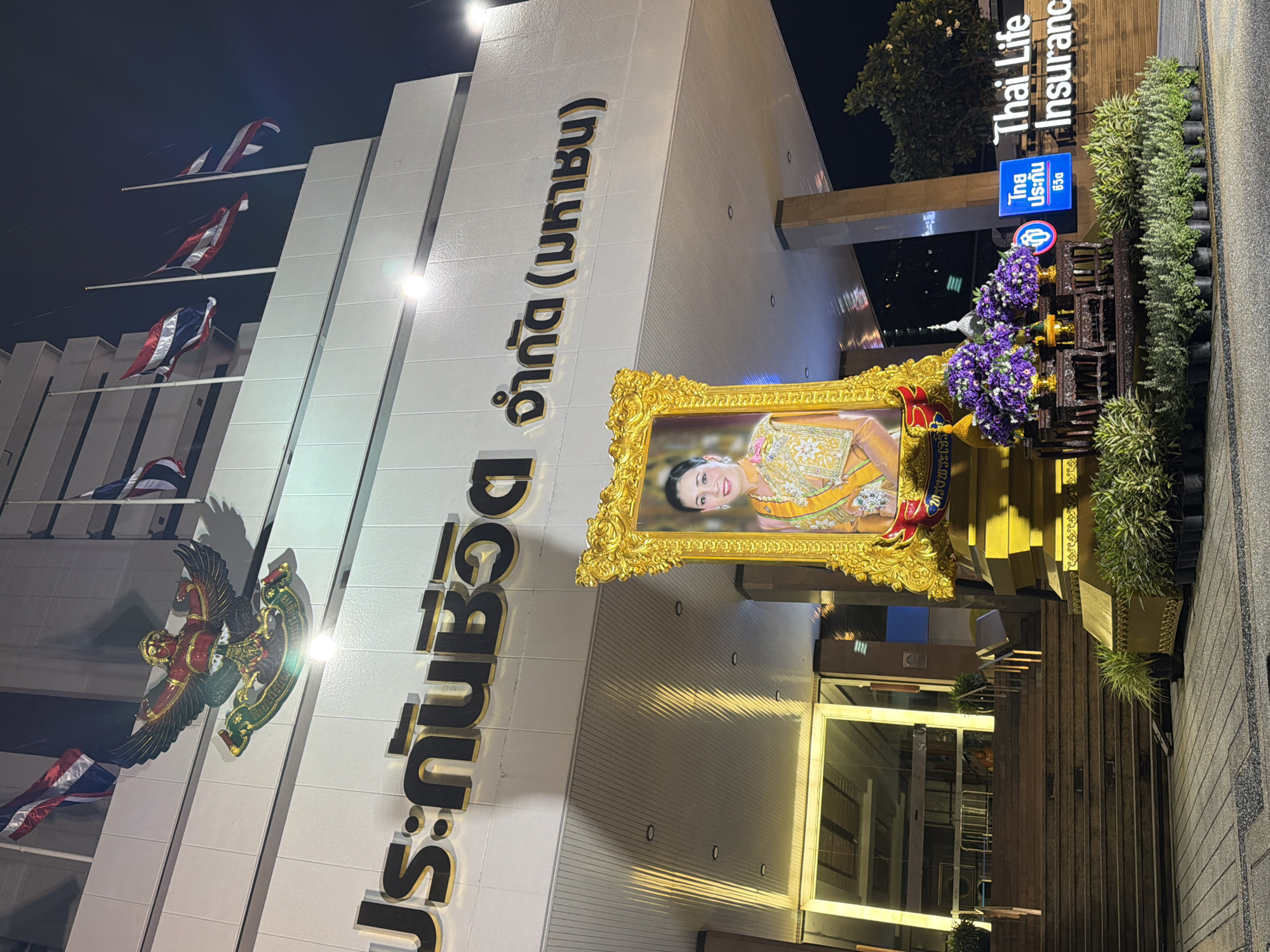 The image shows the entrance of Thai Life Insurance building in Bangkok, Thailand. In front of the building, a large portrait of Queen Sirikit is displayed in a golden frame, adorned with purple flowers and greenery. Thai flags are flown at half-mast on the building, suggesting a period of mourning. The building's facade features the Thai inscription "ประกันชีวิต จำกัด (มหาชน)", which translates to "Life Insurance Public Company Limited". The area is illuminated by artificial lights, creating a nighttime scene.