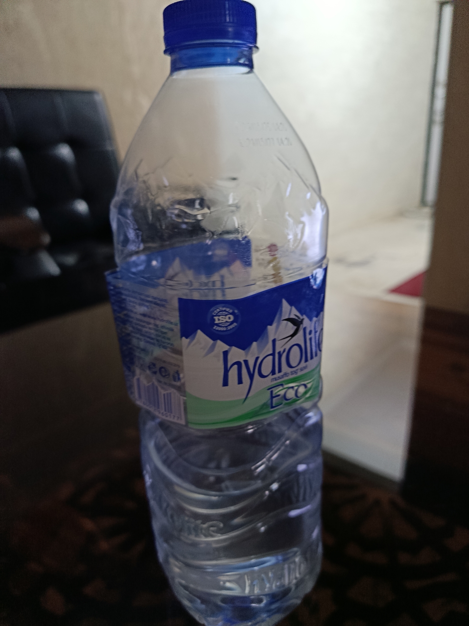 The image shows a plastic bottle of "Hydrolite ECO" water, with a blue cap. The bottle is partially filled with water and is positioned on a dark, patterned surface that appears to be a table or desk. In the background, out of focus, is a black leather chair and a wall with a muted, textured finish. The lighting suggests an indoor setting, likely a room. There are no people or animals present in the image. The location is specified as Tashkent, Uzbekistan, though there are no visual cues within the image to confirm this.
