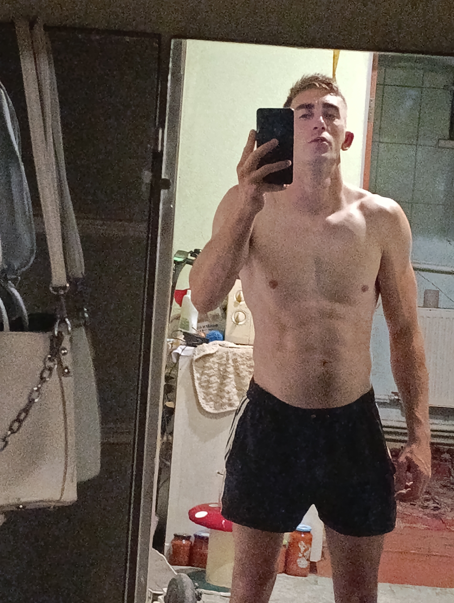 A young adult male with short, styled brown hair is taking a mirror selfie. He is shirtless, wearing black athletic shorts with white stripes on the sides. He has a lean, muscular build. He is holding a black smartphone in his right hand, angled to capture his reflection. The background appears to be an indoor domestic setting, possibly a bathroom or bedroom, with a wardrobe or cabinet visible to the left and a radiator in the background to the right. The lighting is dim, and the image has a grainy quality. There are some items visible on a shelf or counter behind him, including jars and bottles. The location is Fastiv, Ukraine.
