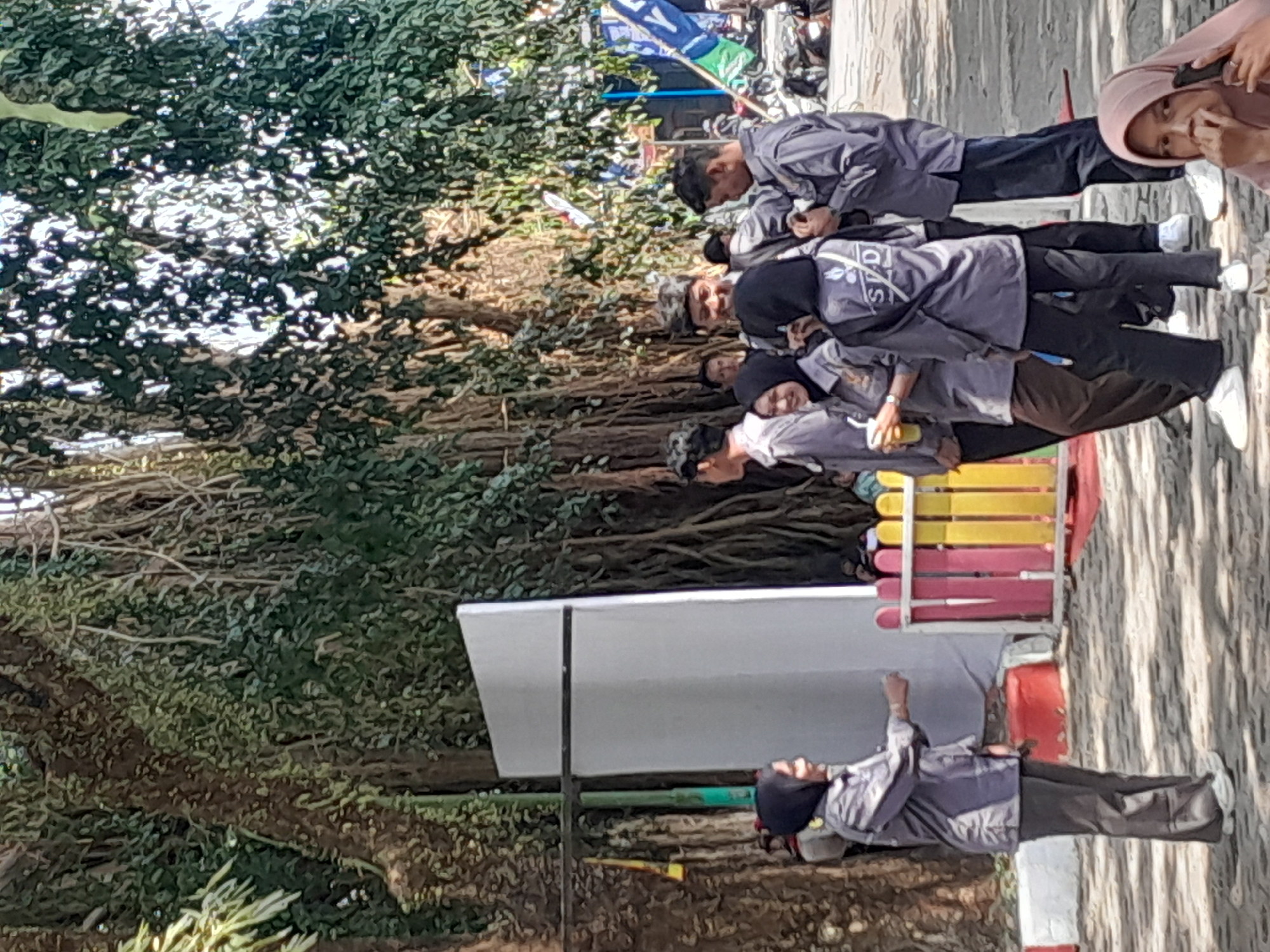 In Genteng Kulon, Indonesia, a group of young people, likely students due to their matching gray uniforms, are gathered outdoors. A large, old tree with a thick, gnarled trunk and abundant foliage dominates the background, casting dappled shadows. In the foreground, several individuals are standing and interacting. One young woman in a hijab and a gray uniform is gesturing with her right hand. Another young woman, also in a hijab and uniform, holds a yellow cup. Two young men, one in a gray uniform and another in a similar shirt with a patterned head covering, are also present. A large white board stands to the left of the group, with a colorful, low fence in front of it. The ground appears to be a mixture of paved and sandy areas. The overall scene suggests a casual outdoor gathering or event.