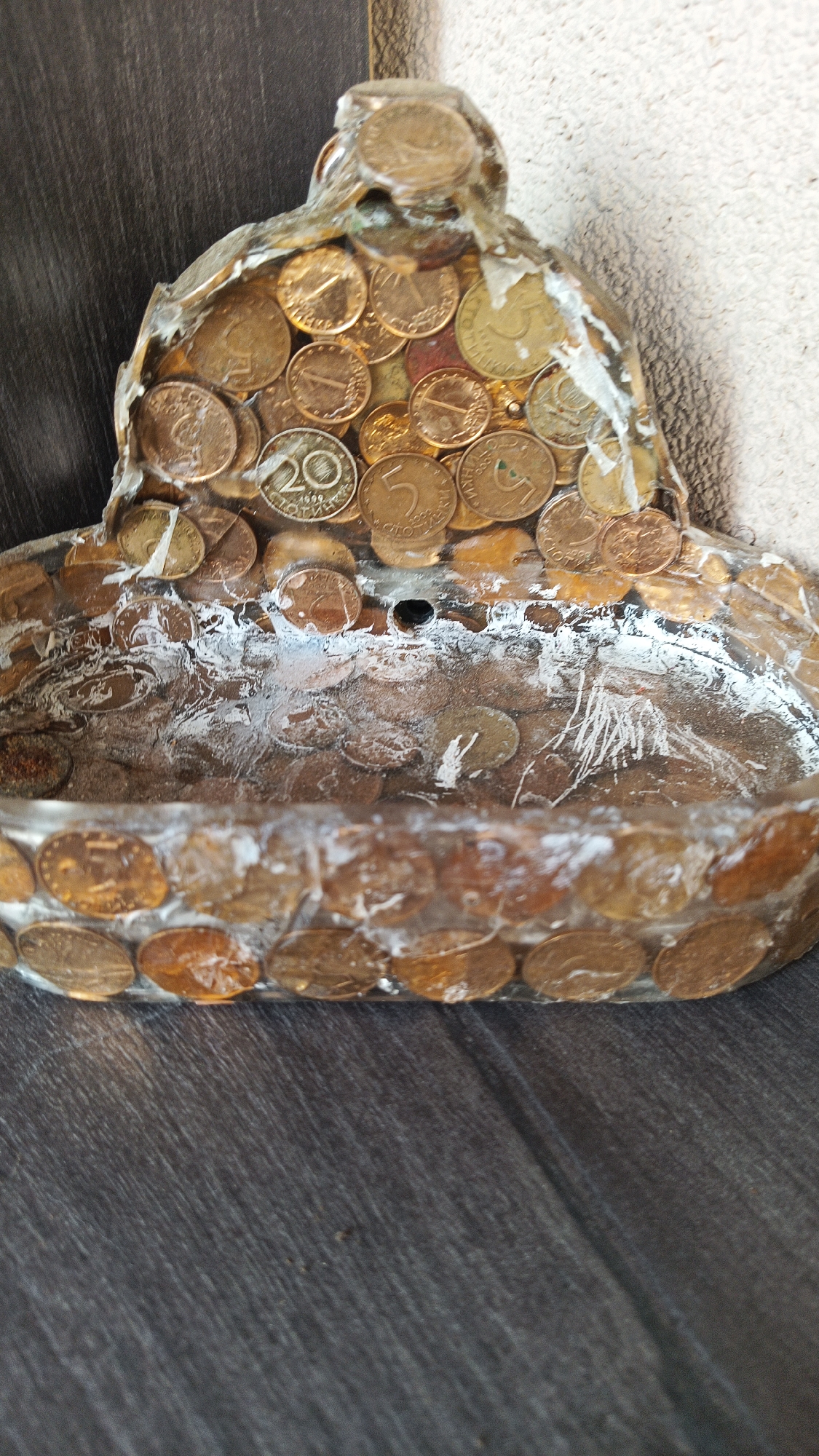 A handmade, coin-covered receptacle sits on a dark, wood-grained surface. The vessel is shaped like a shallow bowl with a raised back. The bowl is lined and rimmed with coins of various sizes, potentially Bulgarian coins, and encased in a clear, resin-like material. White residue or substance is present within the bowl's interior. The raised back of the receptacle is also covered in coins. The background is a dark wooden surface to the left and a neutral beige wall on the upper right. The location might be connected to Karnobat, Bulgaria, based on the presence of what seems to be Bulgarian currency.