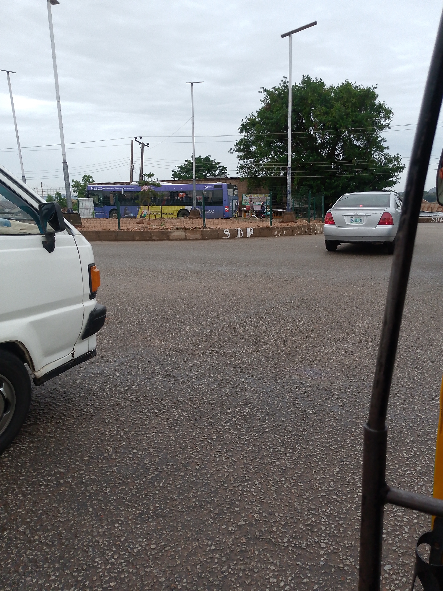 The image depicts a street scene in an urban environment in the city of Unknown, Unknown. A white van is visible on the left side of the frame. A silver sedan is positioned at a roundabout, with its license plate, MKA 801 AL, visible. In the background, a bus with "NESCO" branding can be seen parked alongside a building. Tall street lights are spaced along the road. The sky appears overcast. The foreground shows the asphalt road surface. In the bottom right corner, parts of a three-wheeled vehicle (possibly a tricycle or auto-rickshaw) are visible. The letters "SDP" are painted on the curb of the roundabout.