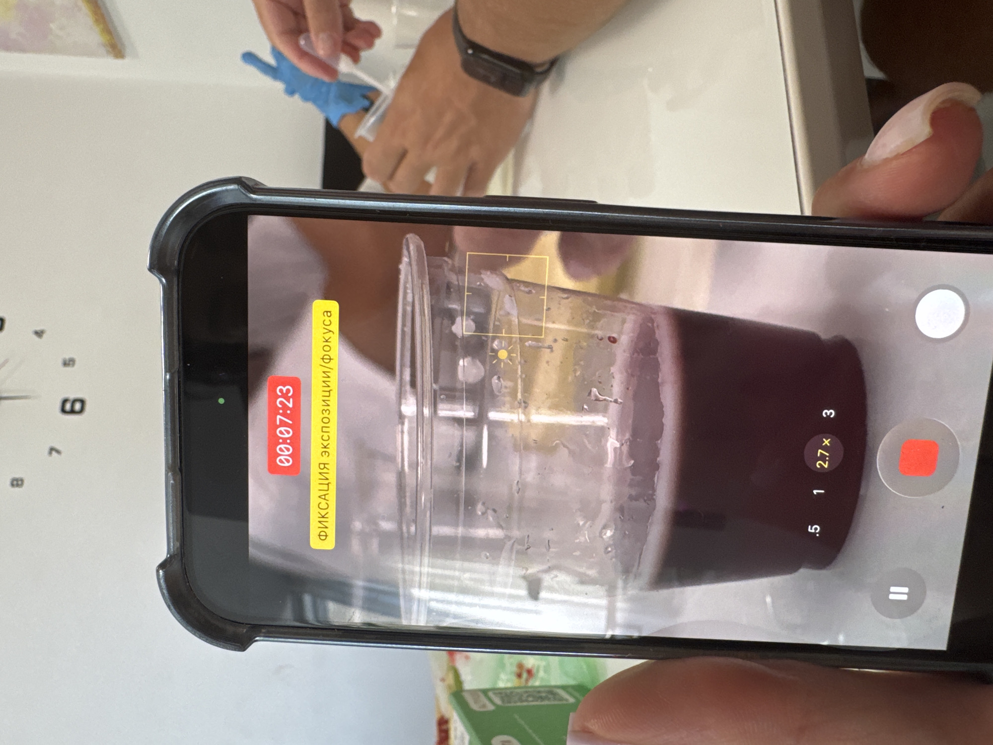 The image shows a close-up of a smartphone screen displaying a video recording. On the screen, a plastic cup filled with a dark purple liquid is visible. The cup has condensation on the inside. Above the cup, a yellow banner displays Russian text that reads "ФИКСАЦИЯ ЭКСПОЗИЦИИ/ФОКУСА," which translates to "Fixation of exposure/focus." A red timer at the top of the screen shows "00:07:23." In the lower part of the screen, there are video controls, including a pause button and a red record button. A zoom indicator shows "2.7x."
In the background, visible over the smartphone screen, are elements of a room. A clock face is mounted on a white wall, with hands pointing to approximately 1:50. To the right, a person wearing blue medical gloves is holding a syringe and a small vial. Another person's arm, wearing a dark smartwatch, is also visible. The overall setting appears to be indoors, possibly a laboratory or medical setting. There is no information in the image that can be used to determine its location in Netanya, Israel.