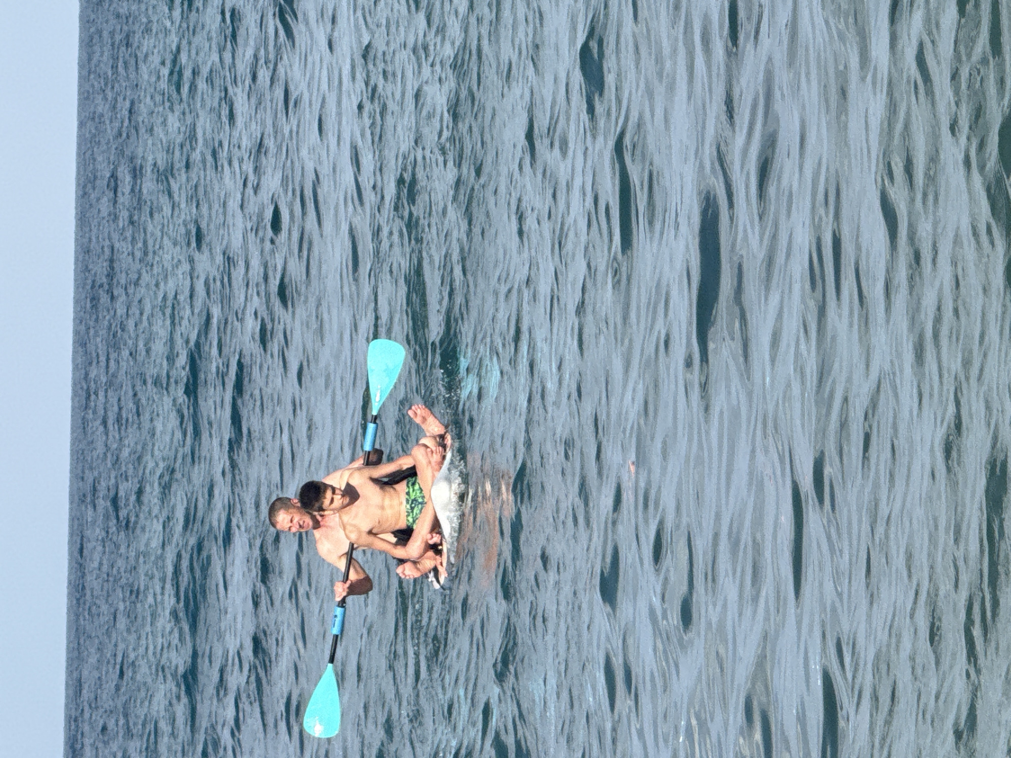 Two individuals, an adult male and a youth, are seated on a paddleboard in the sea. The adult male is positioned behind the youth, holding a paddle that extends across both their bodies. The youth is facing forward, with his legs crossed. Both are shirtless. The paddleboard is white and appears to be floating on the surface of the blue sea. The water is slightly rippled under a clear sky. The image was taken off the coast of Partenit, Ukraine.