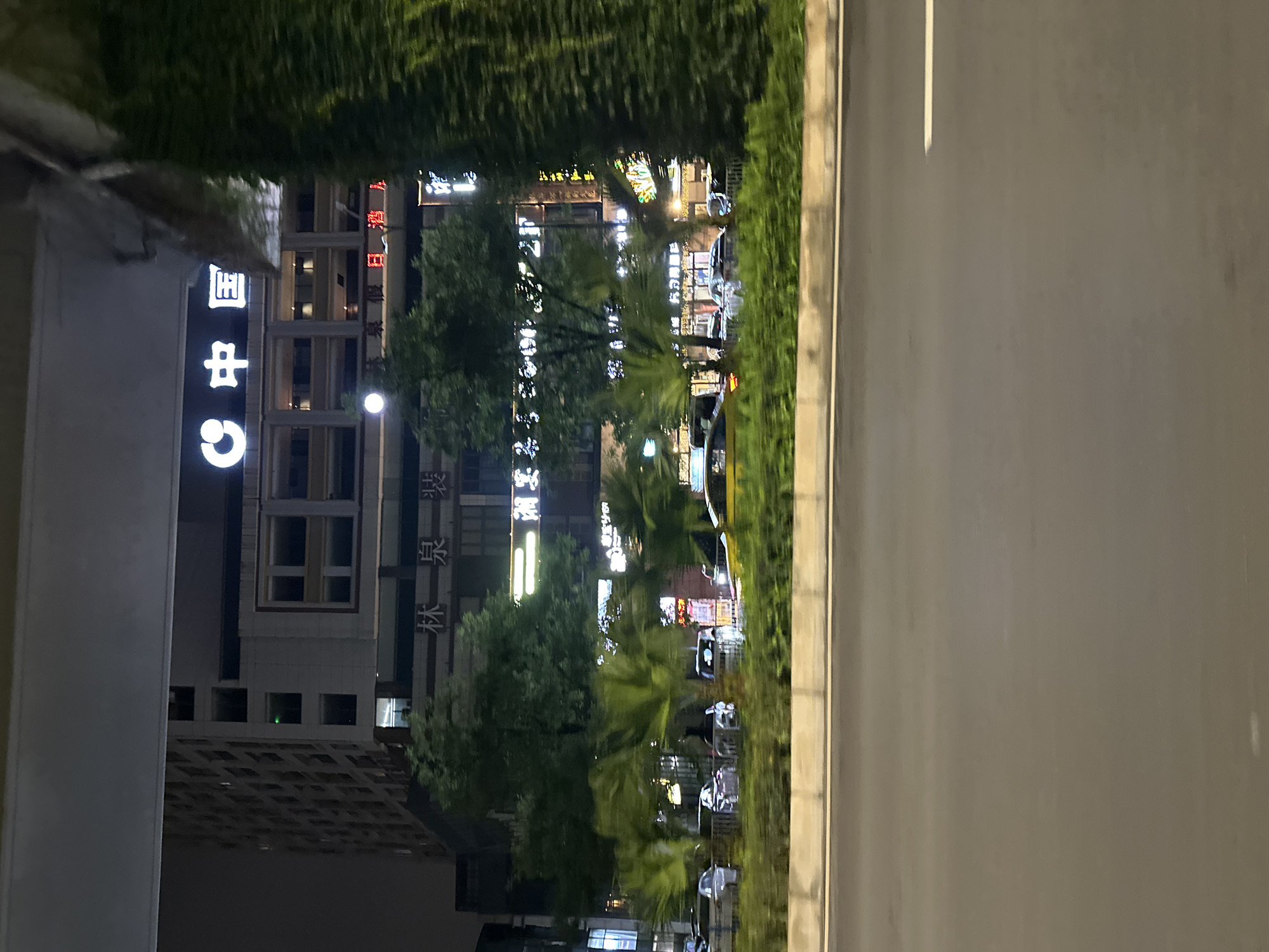 The image captures a nighttime view of a street in Chongqing High-tech Zone, China. A tall, modern building with illuminated signage stands prominently in the background. The signage features Chinese characters, including "中国" which translates to "China," suggesting it might be a prominent corporate building or hotel. Below the main signage, there are more illuminated signs in Chinese, some appearing to be for businesses or services.
In the middle ground, a row of trees, including some palm-like varieties, line the street. Several cars are visible on the road, some parked and some in motion. One yellow vehicle, possibly a taxi, is in the foreground, indicating active traffic. The road surface appears to be asphalt, and a concrete barrier with some greenery on top separates the road from the sidewalk or median.
To the right of the image, a tall, dense structure covered in green foliage, likely a building or a decorative structure, partially obscures the view of the main building and the sky. The lighting suggests a typical urban environment at night, with artificial lights from buildings and vehicles illuminating the scene. The overall impression is that of a busy commercial or residential area within a modern Chinese city.