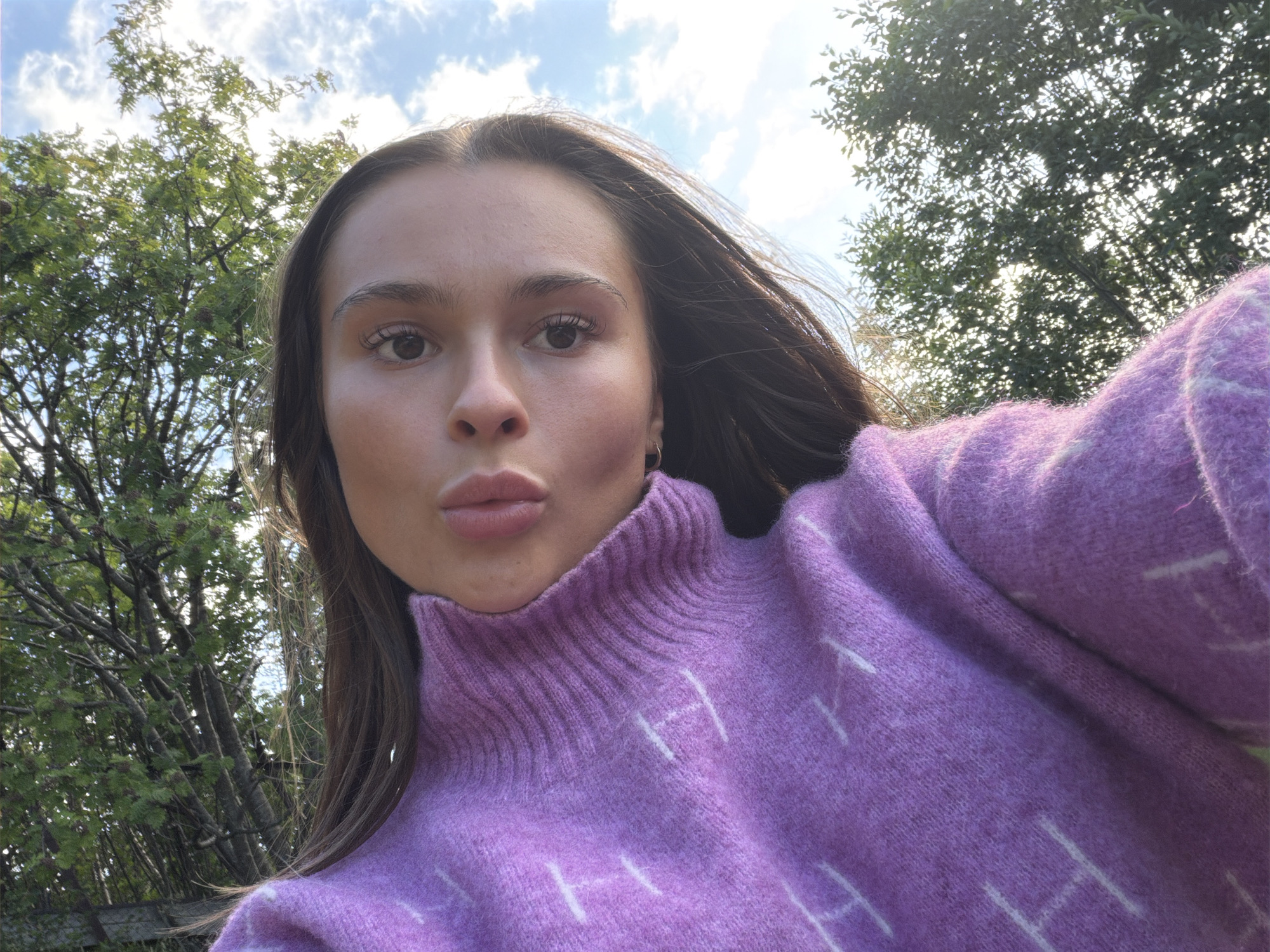 A young woman with long, dark brown hair is captured in a close-up, selfie-style photograph. She is wearing a light purple, knitted sweater with a high neckline and a subtle white pattern. The woman's face is turned slightly towards the camera, with her eyes looking forward. Her lips are slightly parted, and she has natural-looking makeup on. The background consists of lush green trees and a bright blue sky with scattered white clouds, suggesting an outdoor setting. The lighting indicates it is daytime, likely with the sun casting a warm glow. The image does not provide any specific landmarks or context to place it within the city of Harstad, Norway.
