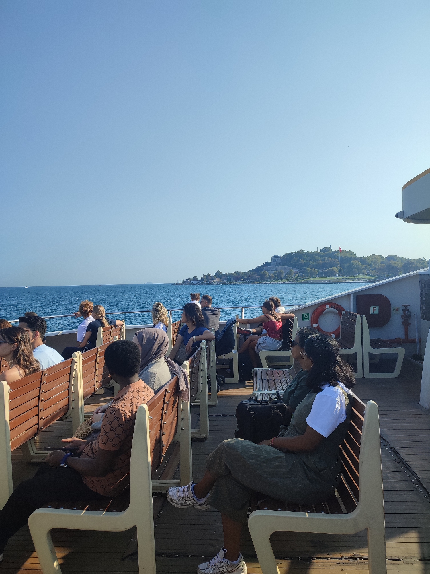 This image depicts a group of people seated on a ferry on a clear, sunny day, with a coastline visible in the distance. The ferry appears to be in the Bosphorus Strait, likely in Istanbul, Turkey, given the architectural style of the distant buildings, which resemble historical structures. The people are diverse in age and ethnicity, dressed in casual summer clothing. They are sitting on wooden benches with white frames, facing either the water or the interior of the ferry. Some individuals are looking out at the sea, while others are engaged in conversation or looking at their phones.
The water is a deep blue, with gentle waves. The coastline is lined with lush green trees and scattered buildings, including a prominent structure with a dome on a hill, possibly a mosque or a historical landmark. The sky is a clear, bright blue, with no clouds visible. The ferry itself has a wooden deck and white seating areas. There are safety railings along the edge of the deck and some safety equipment, such as a life preserver, are visible. The lighting suggests it is daytime, likely late morning or early afternoon, with the sun casting shadows on the deck.