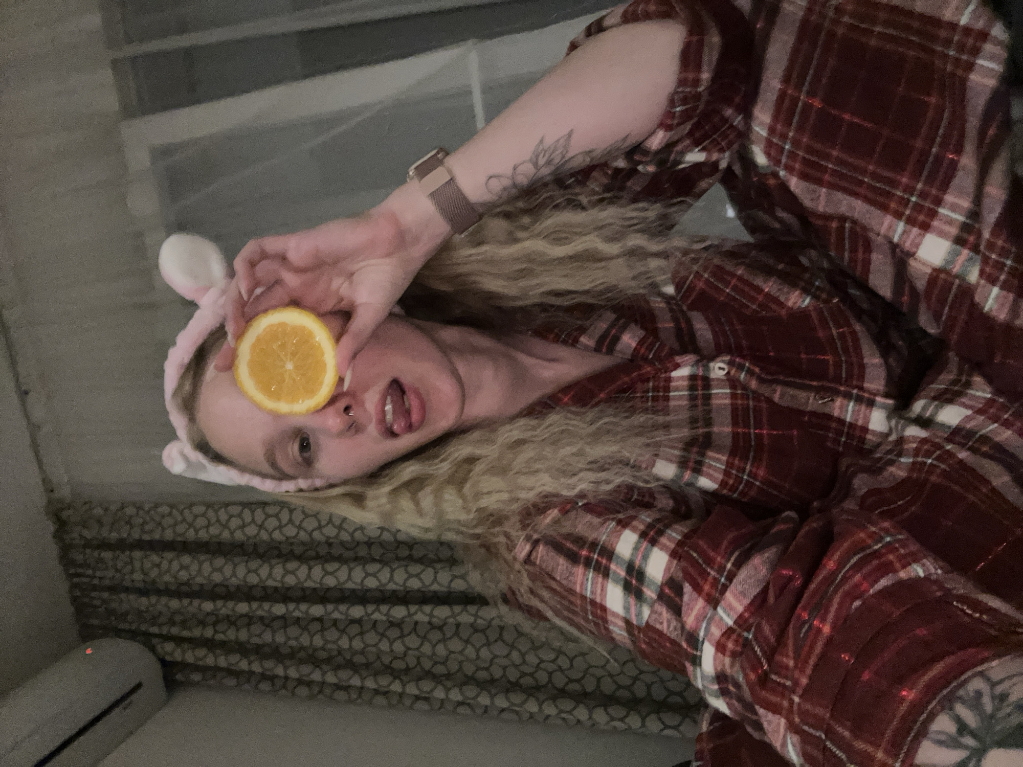 A blonde woman is posing with a slice of orange covering her eye. She is wearing a pink headband with bunny ears and a red and white plaid shirt. She has tattoos on her arms and a watch on her left wrist. Her tongue is sticking out. The background shows a curtained window and what appears to be an air conditioning unit. The location cannot be confirmed as Budva, Montenegro, based solely on this image.