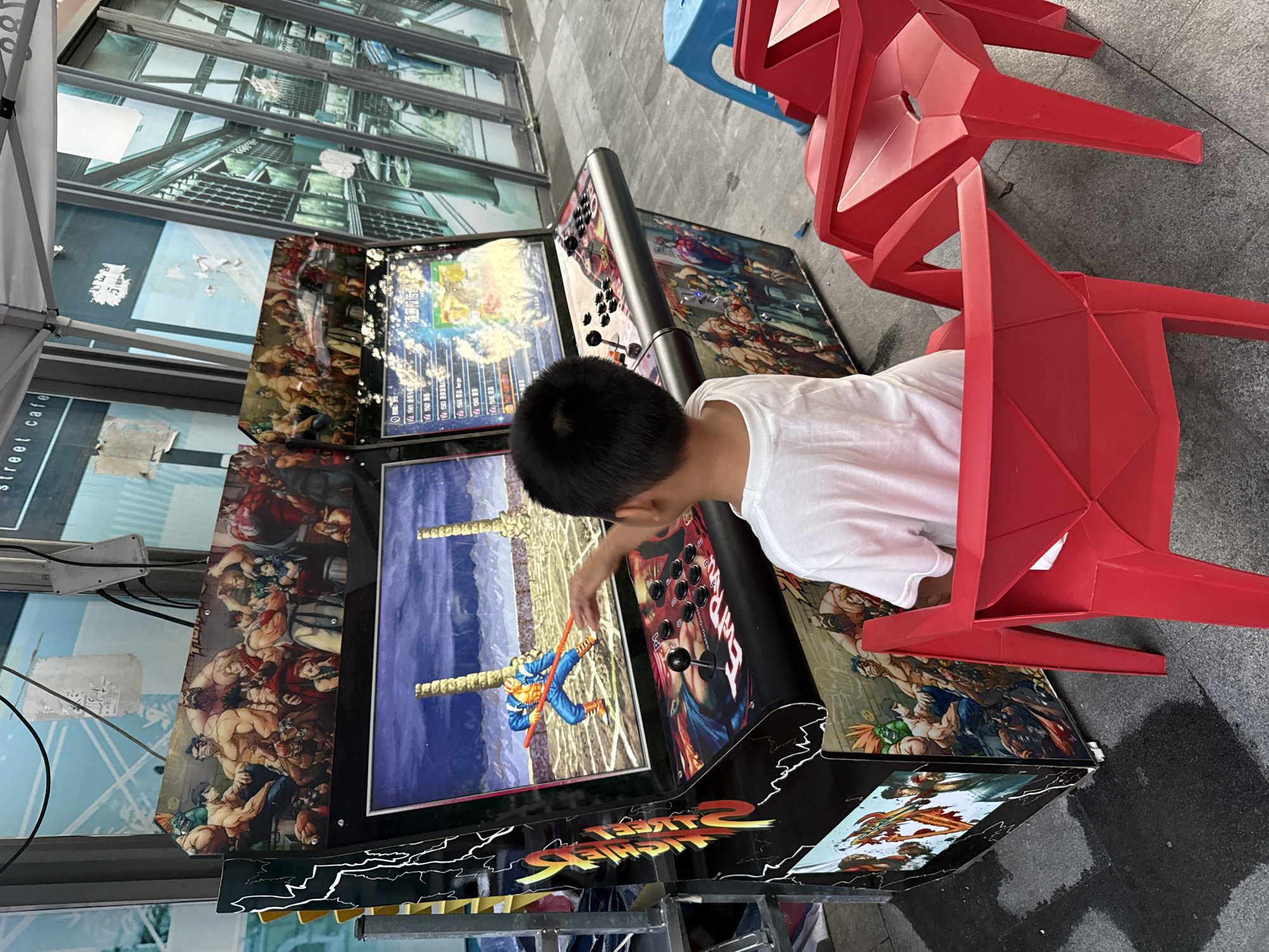 A young boy in a white t-shirt is playing a vintage arcade game. The arcade machine features artwork from the fighting game "Street Fighter." The boy is positioned on a red plastic chair and his right hand is extended towards the screen, as if he is interacting with the game. The arcade machine has a black body decorated with lightning bolt graphics and characters from the game. The screen displays a fighting scene with characters and game elements. Two other red plastic chairs are visible to the right of the boy. The arcade machine and chairs are placed on a paved surface, possibly a sidewalk or plaza, in front of a building with large glass windows. The background shows a street cafe sign and reflections in the glass, suggesting an urban environment. The scene appears to be outdoors, possibly under a tent or awning. The overall atmosphere suggests a casual entertainment setting.