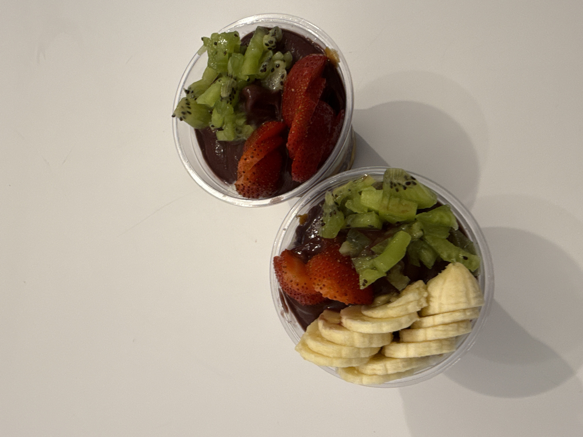 Two clear plastic cups filled with a dark, smoothie-like substance are arranged diagonally on a white surface. The cup in the foreground is closer to the viewer and positioned to the right. This cup is topped with sliced bananas, forming a semi-circular arrangement along the right side, and chopped kiwi fruit and sliced strawberries on the left. The cup in the background, slightly further away and to the upper left, is also topped with chopped kiwi and sliced strawberries. The lighting suggests an indoor setting, possibly with natural light casting shadows to the right of the cups. The overall impression is that of a healthy, refreshing dessert or snack. There is no visible text in the image.