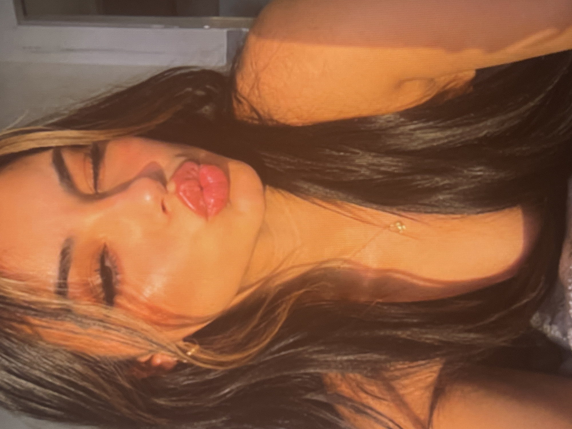 A young woman with long, dark hair and highlights is shown from the chest up, with a bright orange light illuminating her face and upper body. She is leaning to her left, with her right eye closed and her lips puckered in a kiss. She is wearing a simple necklace with a small pendant. Her left shoulder is visible, and her right arm is slightly bent and resting at her side. The background is a soft blur of light and shadow, suggesting an indoor setting.