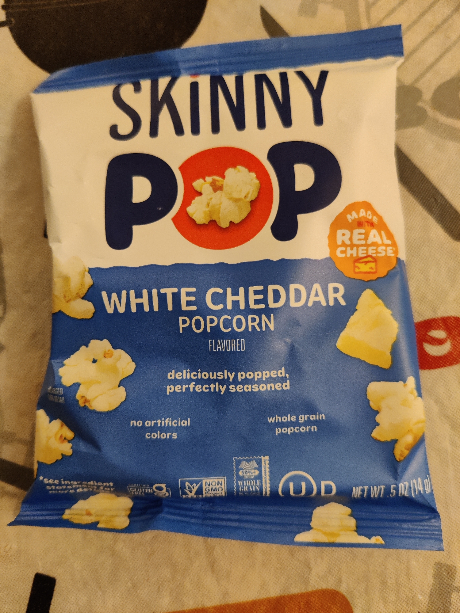The image shows a bag of Skinny Pop White Cheddar Popcorn. The bag is predominantly blue with white and orange text and graphics. The brand name "SKINNY POP" is prominently displayed in large, dark blue lettering at the top. Below this, the words "WHITE CHEDDAR POPCORN" are written in white. Various images of popcorn kernels and pieces are scattered across the bag. A circular orange label indicates "MADE WITH REAL CHEESE". At the bottom of the bag, nutritional information and certifications such as "NON GMO Project VERIFIED", "CERTIFIED GLUTEN FREE", and a "50%+ WHOLE GRAIN" seal are visible, along with the net weight ".5 OZ (14g)". The bag is placed on a surface that appears to be a tablecloth with a subtle pattern. The lighting suggests an indoor environment. There are no people or animals present. The specific location in Lower Paxton Township, United States of America, is not discernible from the image alone.