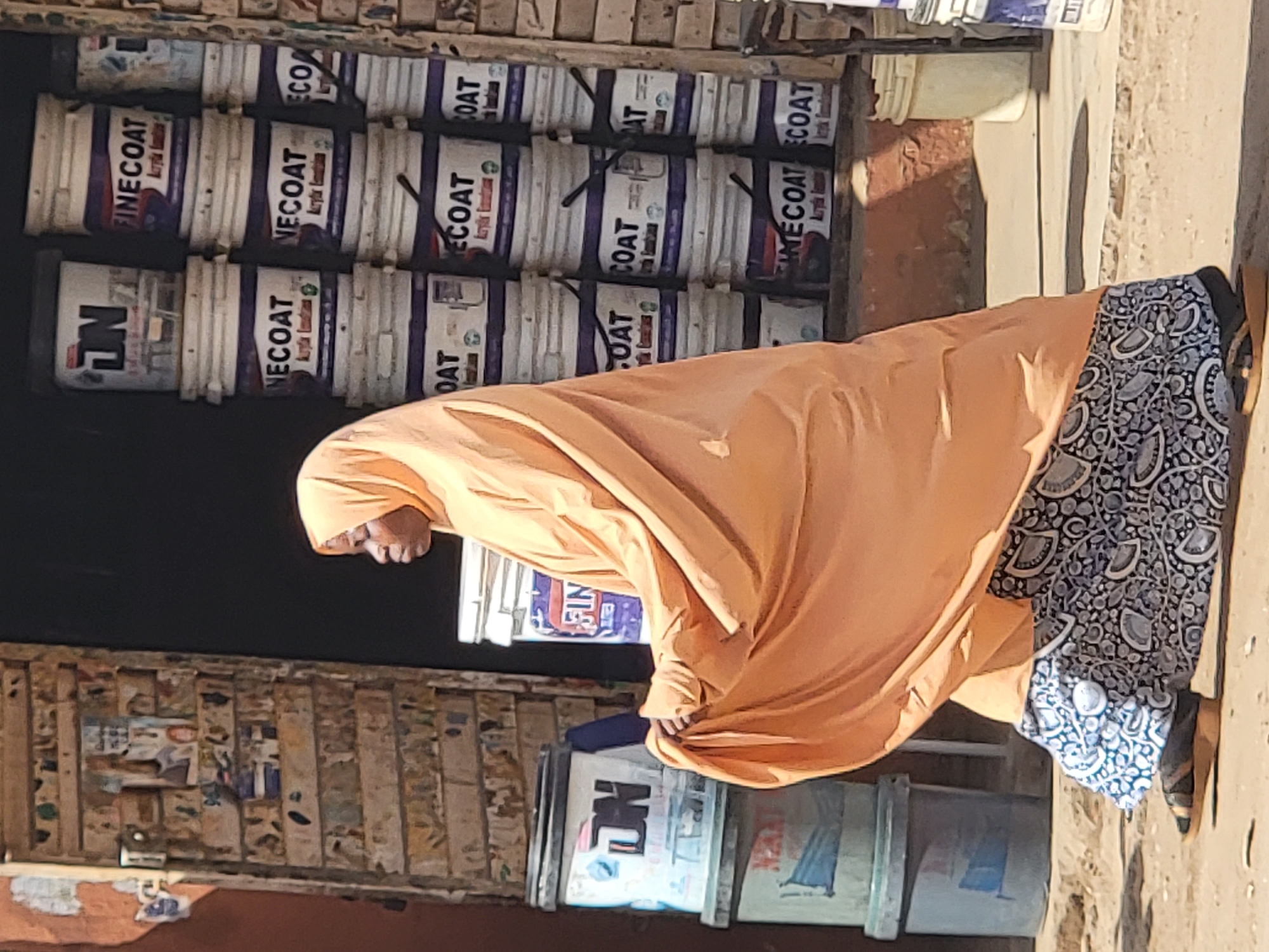 A woman in an orange headscarf and blue patterned dress walks along a street in Maiburiji, Nigeria. Behind her, stacks of "FINECOAT" and "NECOAT" paint buckets are visible, as well as a wooden structure with posters or advertisements attached to it. The environment appears dusty and sunny.