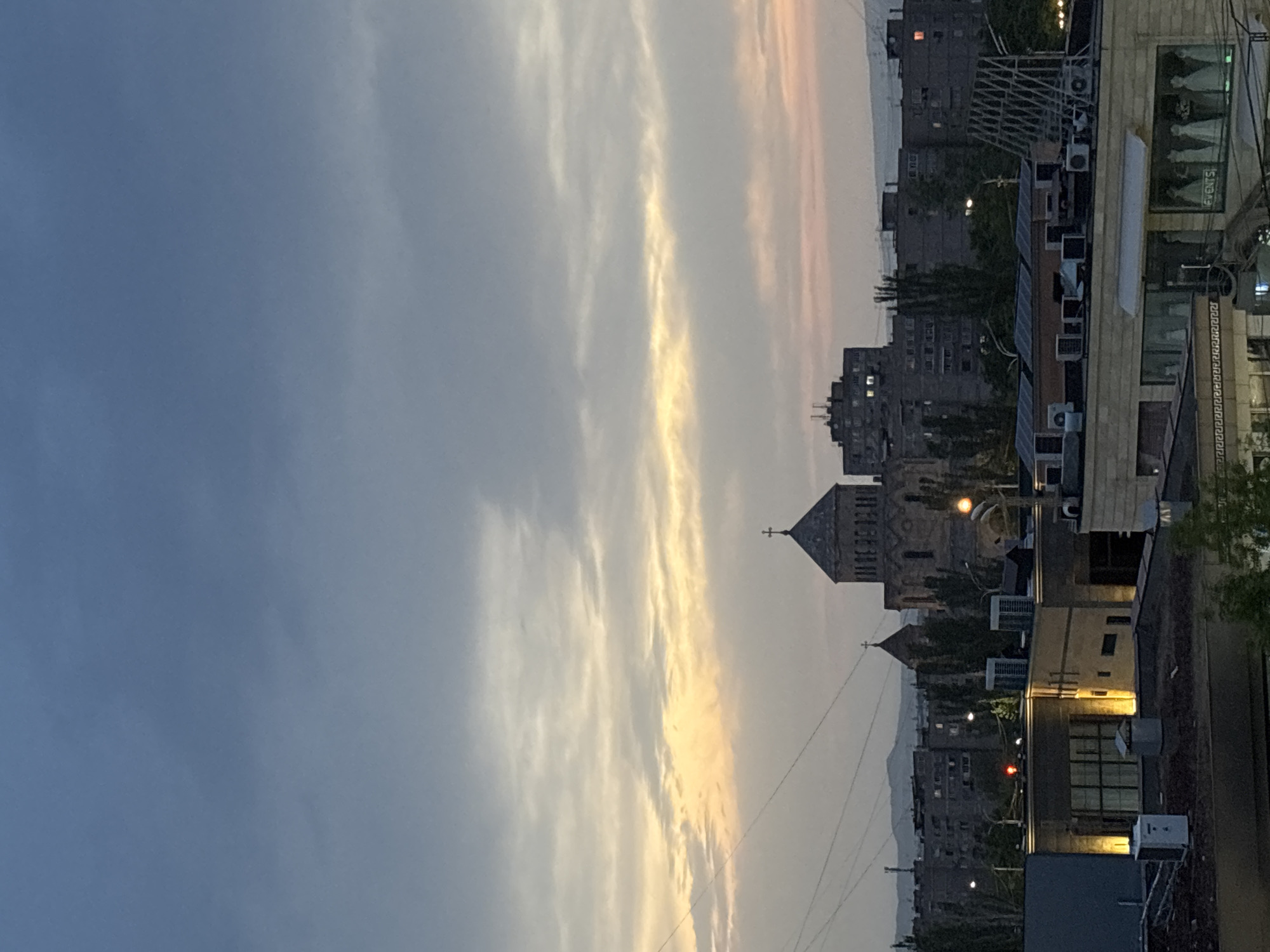 The image depicts a view in Yerevan, Armenia at twilight. In the middle of the frame, there is a church with pointed towers. A row of buildings extends from the right edge, some with visible air conditioning units and reflective windows. Beyond the church and buildings are urban apartment structures. The sky is primarily blue with streaks of cloud formations that are highlighted by golden light from the setting sun.