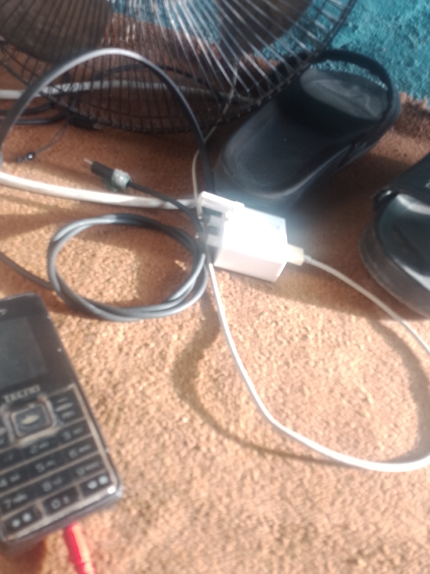The image shows a close-up view of electronic devices and personal items on a textured surface, likely a rug or carpet. In the foreground, a black mobile phone with a keypad is visible, with a red charging cable plugged into its port. A coiled black cable lies next to the phone, and a white charging adapter is connected to a white cable that extends out of the frame. In the background, a black sandal is positioned on the surface, and behind it, a portion of a fan's grille and blades can be seen. The lighting suggests an indoor setting, possibly a domestic environment. There is no visible indication of the city of Jalingo, Nigeria.
