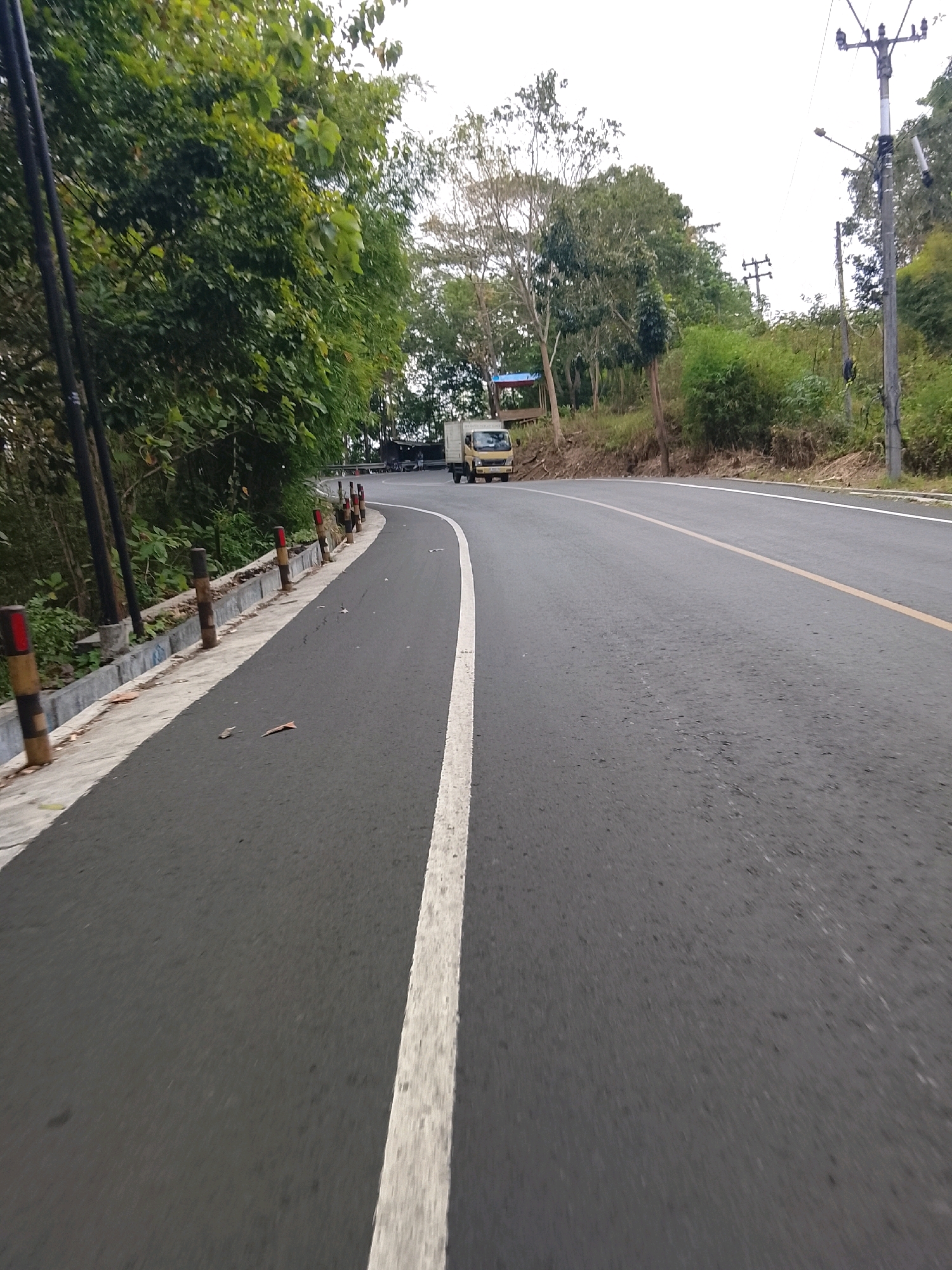 This is a forward-facing, eye-level shot of a paved road curving to the left, situated in Banjar, Indonesia. The road has a white line on the left edge and a yellow line dividing the lanes in the distance. On the left side of the road, there is a sidewalk with a low concrete barrier and wooden posts with red and yellow reflective markers. Lush green trees and foliage line the embankment above the sidewalk, and several utility poles with wires are visible in the background. Further down the road, a small yellow truck is driving away from the camera, and there are signs of a few scattered buildings and structures on the hillside. The sky is overcast and bright.