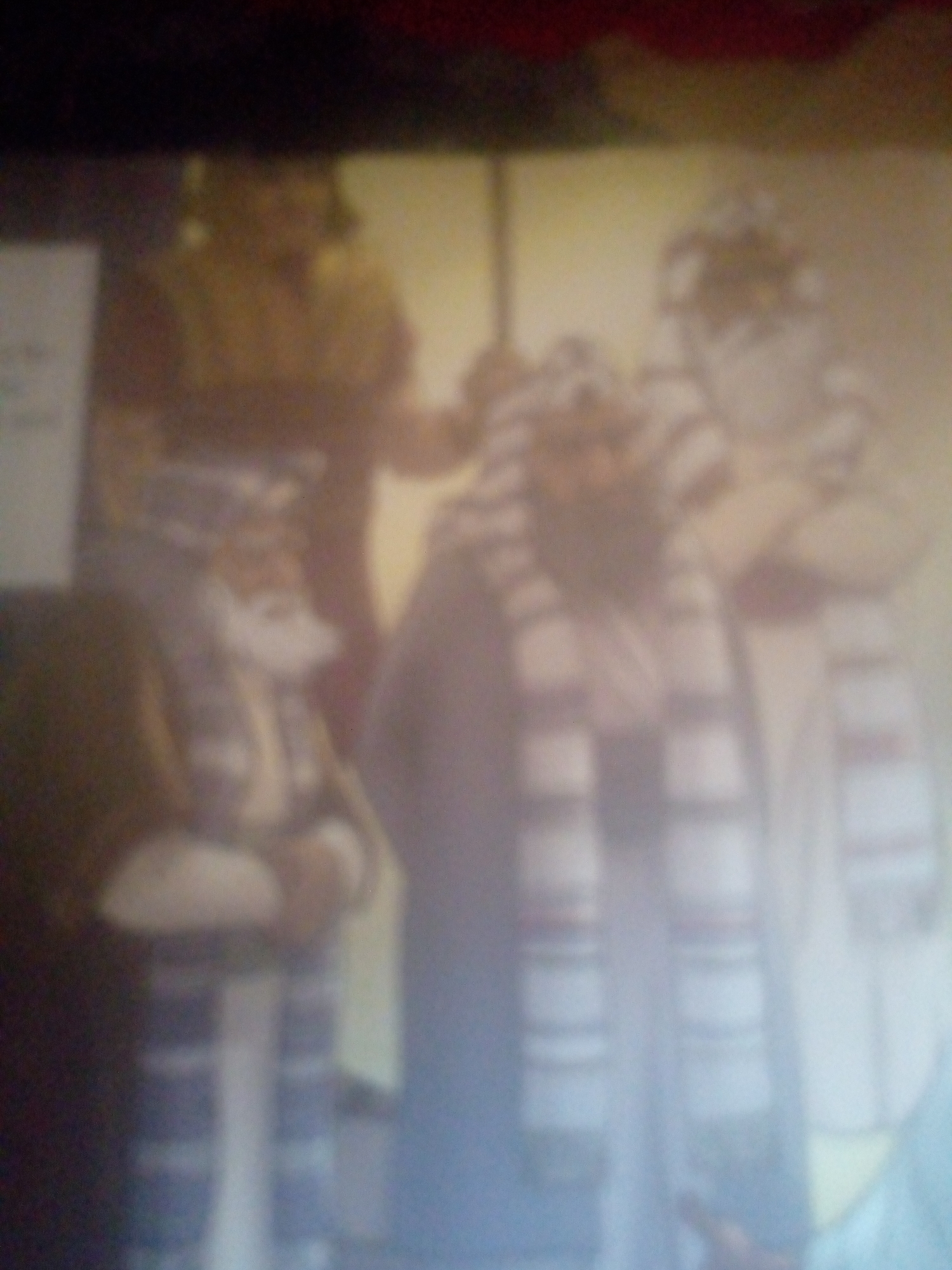 The image shows a group of people in what appears to be a performance or reenactment, possibly for a religious or historical event. There are at least three individuals visible, dressed in robes and head coverings. One individual in the foreground is wearing a white and blue striped scarf or sash. The background is blurred, but suggests an indoor setting with some yellowish lighting. Without further context or clearer details, it is impossible to ascertain the specific location within Jalingo, Nigeria, or the exact nature of the event.