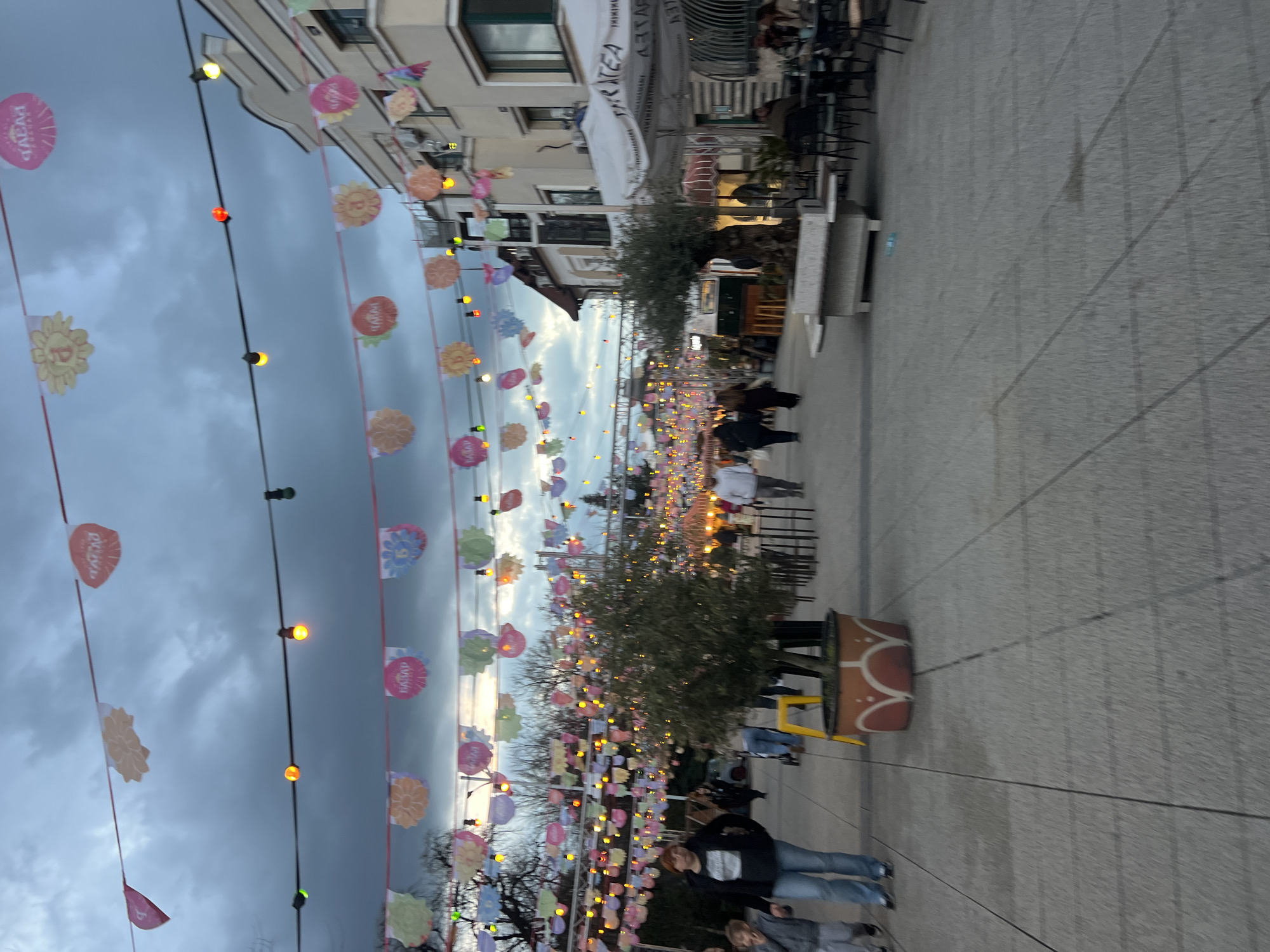 The image depicts a pedestrianized street in Plovdiv, Bulgaria, adorned with strings of decorative flags and lights. People are walking along the street. There are trees and potted plants lining the pavement. Buildings are visible on either side of the street, with some having outdoor seating areas. The sky is overcast.