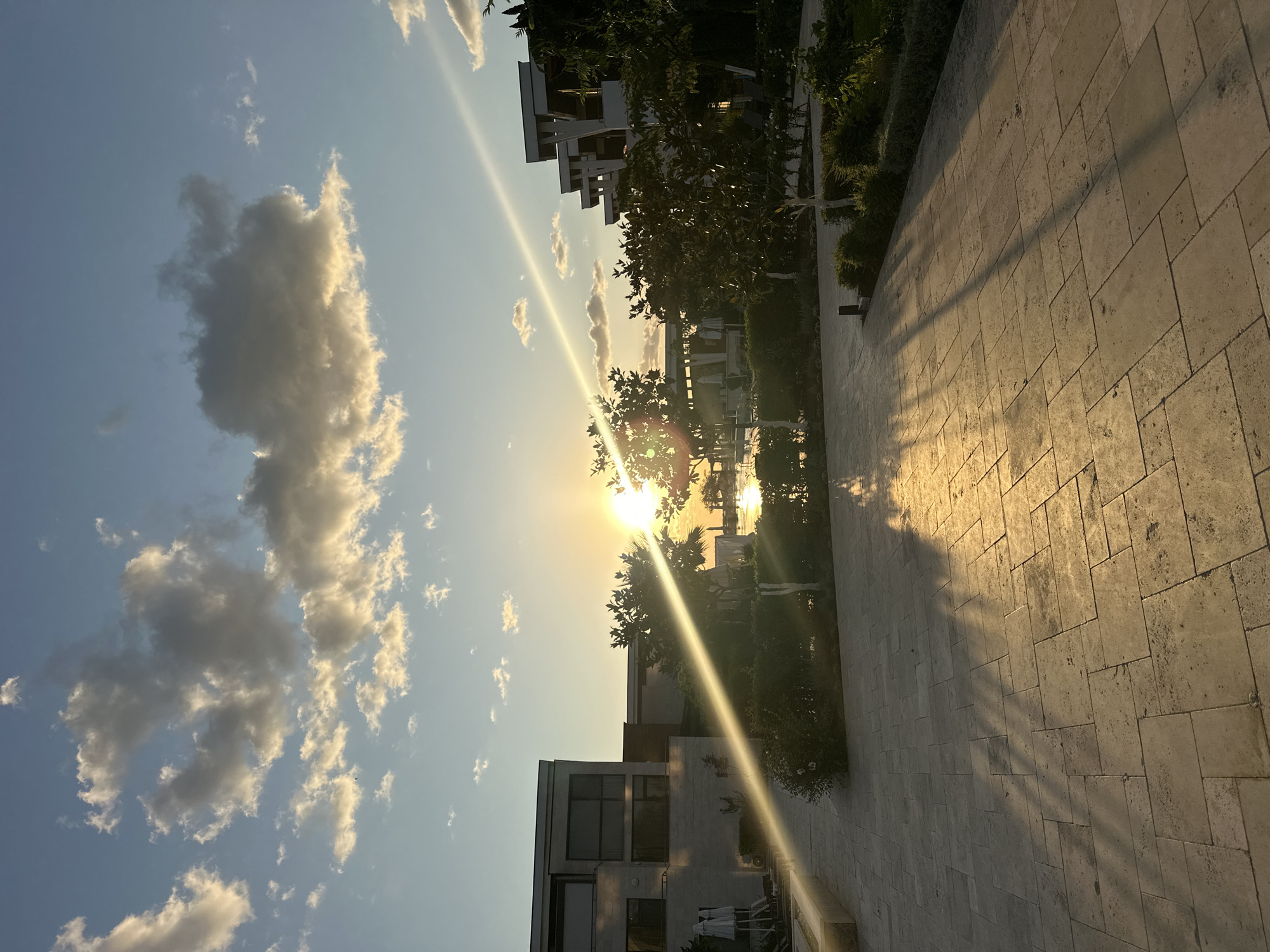 The image depicts a sunset at a modern resort or hotel complex in Sozopol, Bulgaria. The sun is low on the horizon, casting a warm, golden light across the scene and creating long shadows on the paved courtyard. A prominent diagonal sunbeam cuts across the frame, originating from the right side and extending towards the left.
On the left, a multi-story building with large windows and a light-colored facade is visible. In front of it, lounge chairs and umbrellas are arranged near a shallow water feature. To the right, another building with a contemporary architectural style, featuring stacked, angular elements, is partially obscured by lush green trees. The vegetation appears well-maintained, with manicured bushes and trees.
The courtyard itself is paved with large, light-colored rectangular tiles, laid in a staggered pattern. The sky is a clear blue with scattered, fluffy white and grey clouds, illuminated by the setting sun. The overall impression is one of a tranquil, luxurious resort setting at the end of the day. No people are visible in the image.