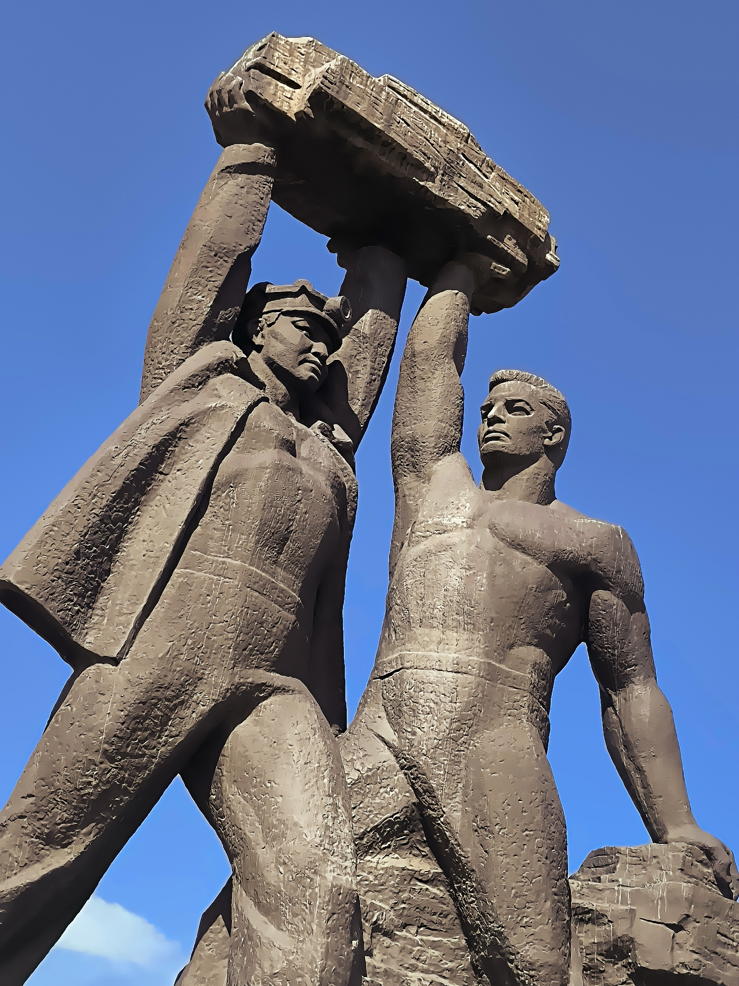 A monumental bronze sculpture depicts two muscular men lifting a large, roughly hewn stone slab against a clear blue sky. The figure on the left wears a cap and a cape that drapes over their shoulder. The figure on the right is bare-chested, revealing a muscular physique. Both figures strain upwards, their arms raised to support the weight of the stone. The sculpture appears to be part of an outdoor monument located in front of the Qarağandı city administration building in Kazakhstan.