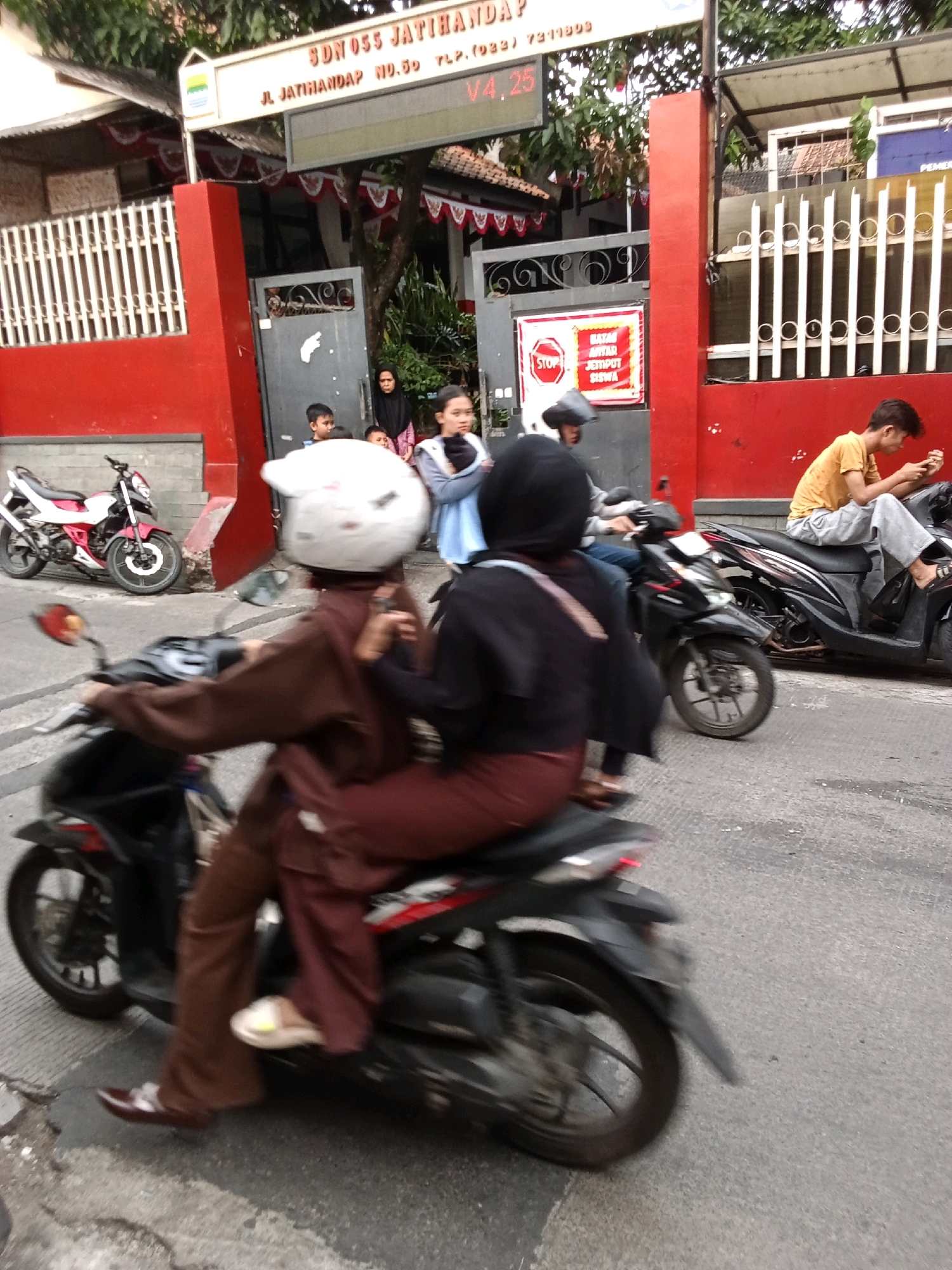 The image depicts a street scene in Bandung City, Indonesia, likely in front of a school given the signage which reads "SDN 055 JATIHAMPRA" and appears to be an address and phone number. In the foreground, two individuals are on a motorcycle. The person in the front is wearing a brown hijab and brown pants and is riding the motorcycle. The person behind them is wearing a black hijab and black top, and appears to be holding onto the rider. Their faces are not clearly visible.
To the right of the foreground motorcycle, another motorcycle is parked, with a young man wearing a yellow t-shirt and grey pants sitting on it and looking at his phone. Behind him, another motorcycle is partially visible.
In the background, there are several people standing near a red building with white railings. Some are children, and some appear to be adults. There are red and white decorations hanging from the building. A sign with a red stop sign and Indonesian text is visible on the wall. The environment appears to be an urban street with a paved road. The overall lighting suggests it is daytime.
