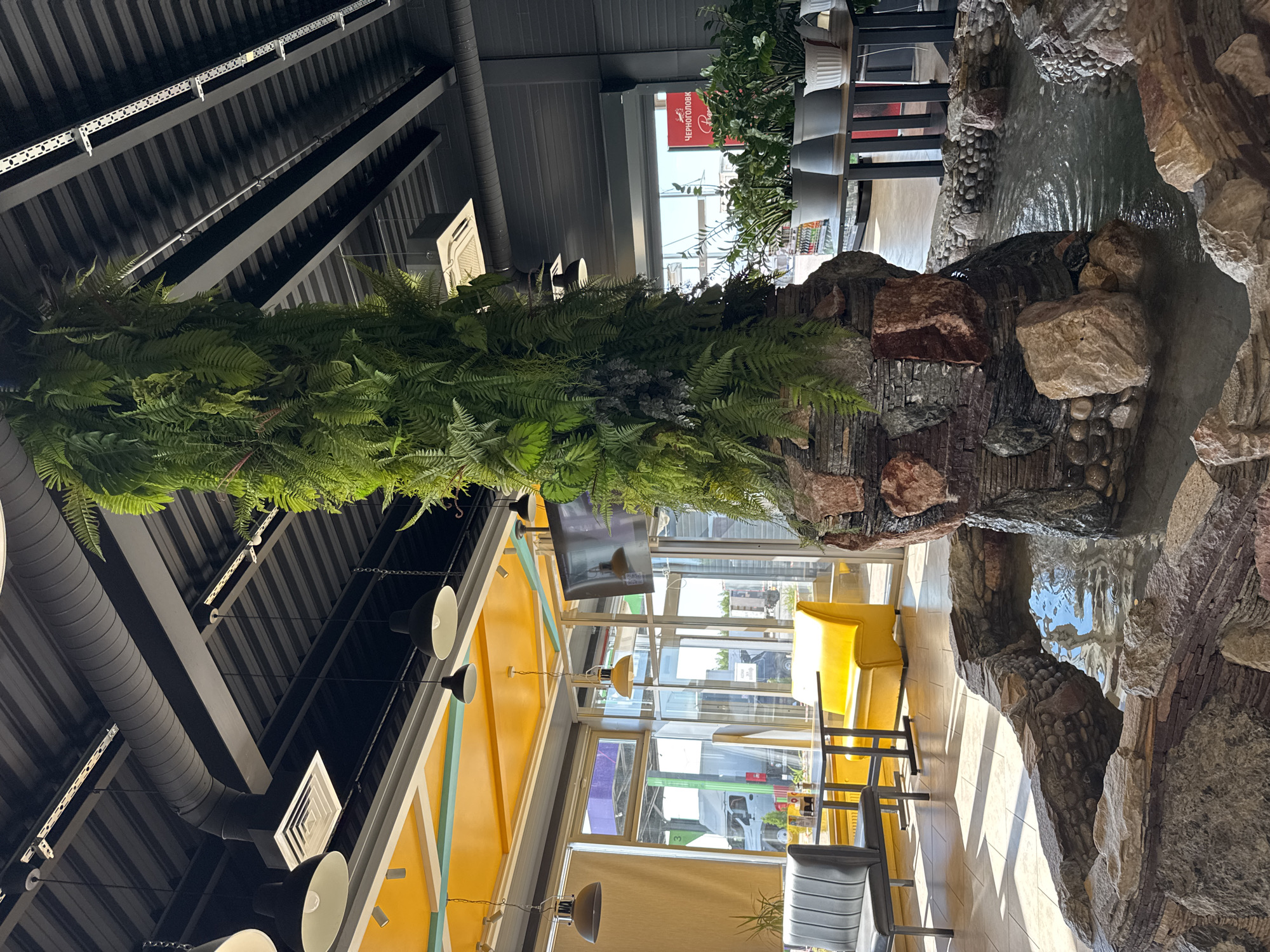 The image captures an interior view within a public space, possibly a restaurant or lounge, located in Казань, Russia. The space is characterized by an industrial-style ceiling with exposed metal beams, ductwork, and hanging light fixtures. A significant feature is the prominent, artificial plant arrangement, likely a fern, suspended from the ceiling, acting as a decorative element.
The setting is oriented towards a bright, glass-paneled wall or window offering views of an exterior scene, featuring vehicles, buildings, and signage suggesting a commercial or urban area. Furniture includes yellow lounge seating and chairs arranged around tables. A decorative water feature constructed with rocks and stonework is also visible. The image appears to be taken from a low angle, highlighting the architectural and decorative elements of the interior space.