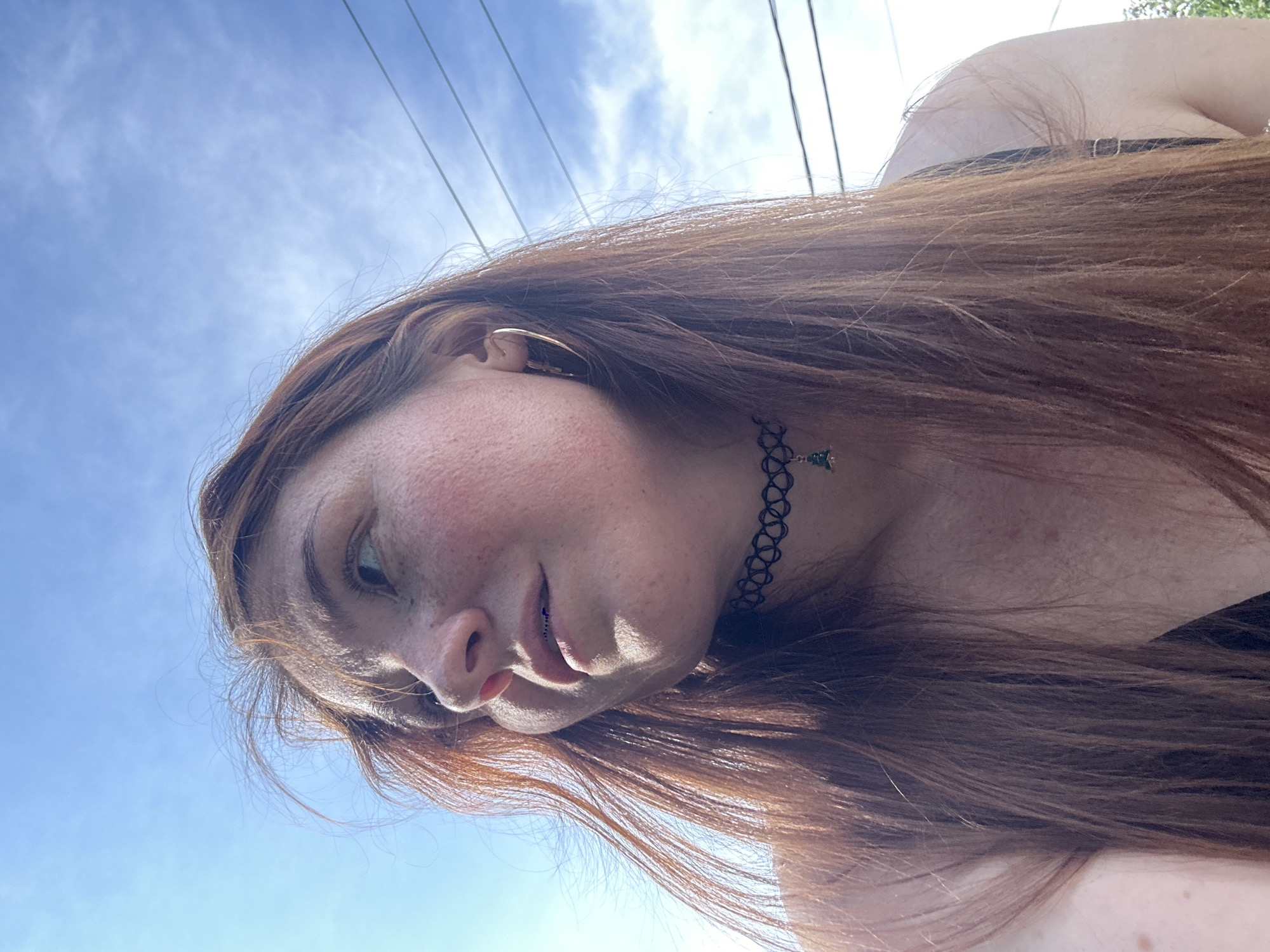 A young woman with long, reddish-brown hair is outdoors, looking up and to the left. She has fair skin with freckles and is wearing a black choker necklace with a small green charm. Her hair is windswept, and she has light-colored eyes and visible braces. The background is a bright blue sky with some wispy clouds and power lines. The setting is likely a residential area of Nizhny Novgorod, Russia, possibly a yard or street, indicated by the presence of power lines and the overall outdoor ambiance. The perspective is from a low angle, looking up at the woman.