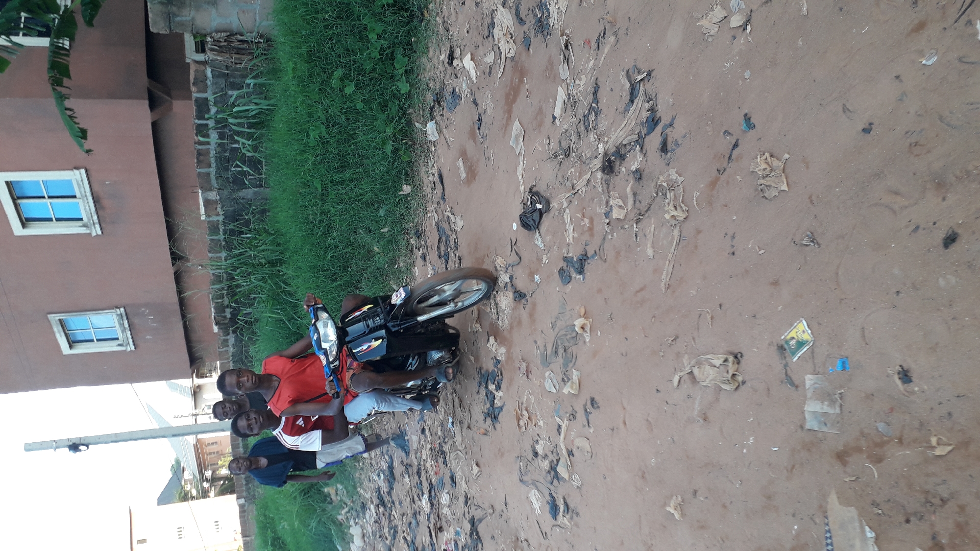 The image shows a group of five young males positioned behind a parked black motorcycle in Nkpor, Nigeria. The setting is a dirt road heavily littered with trash. To the left, there is a building with two visible windows. Adjacent to the road, there is an area of overgrown green grass. The males are casually dressed and appear to be interacting with the motorcycle.
