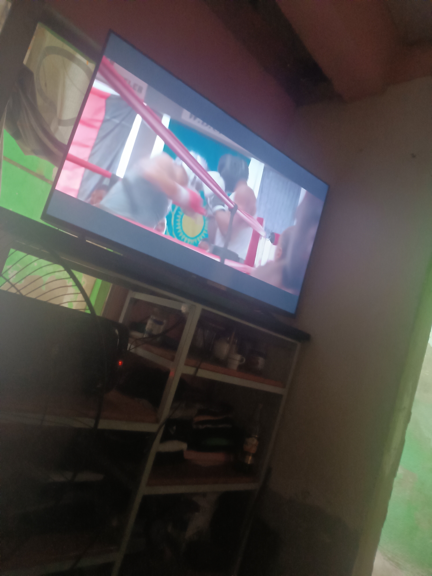 The image shows a television set displaying a boxing match, likely set in a domestic environment. The TV is placed on a multi-tiered shelving unit containing various objects including dishes, clothing, and decorative items. A fan is also visible near the television. The room appears to be dimly lit with light entering through a window in the background. The precise location within the city of Porlamar, Venezuela cannot be determined from the image alone.