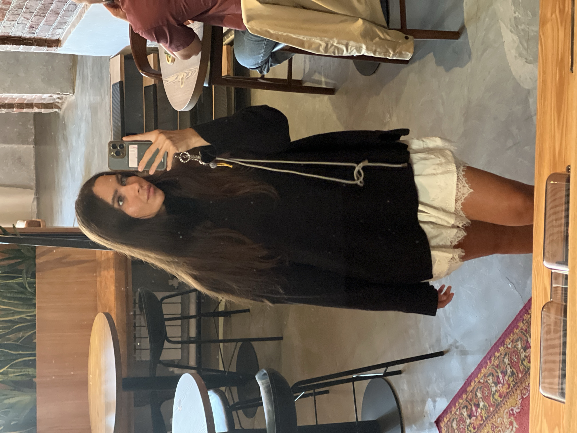 The image appears to be a selfie taken in an interior space that resembles a cafe or restaurant.
People:
*A woman is holding a smartphone to take the picture.
*Another person is partially visible in the background, seated at a table.
Subjects:
*The focus is on the woman in the selfie.
*Clothing: The woman is wearing a black sweater or jacket over a white dress with lace trim.
*Hair: The woman has long, dark hair with a blonde streak in the front.
Objects:
*Smartphone: The woman is holding a smartphone with a wristlet attachment.
*Tables and Chairs: There are wooden tables and metal chairs visible in the background, suggesting a cafe or restaurant setting.
*A rug is located on the lower right of the scene.
Environment:
*The background suggests an interior space, possibly a cafe or restaurant.
*Lighting seems moderate and natural.