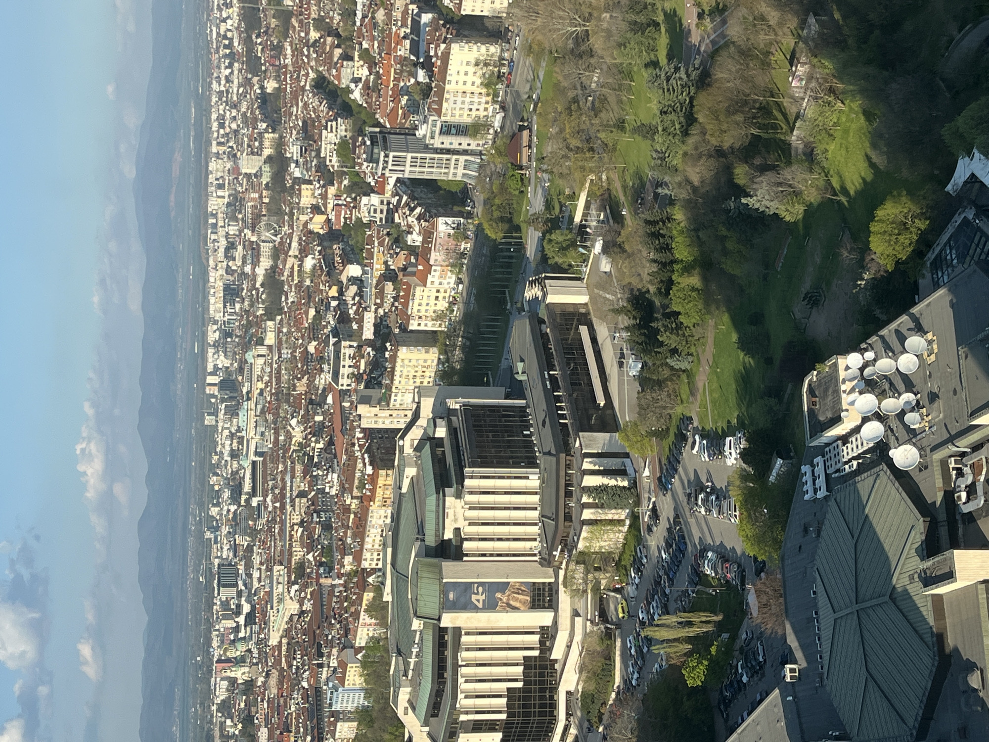This is an aerial view of Sofia, Bulgaria, looking out over the city towards the mountains in the distance. The foreground is dominated by the rooftops of a modern building, featuring multiple satellite dishes. To the left and down the image, there are other modern buildings, including one with a large advertisement. A tree-lined road and a parking lot filled with cars are positioned adjacent to the buildings. Beyond the buildings, the city sprawls out with a dense arrangement of buildings, predominantly with red-tiled roofs. In the background, mountains are visible under a blue sky with some clouds. The scene appears to be taken during daylight hours.