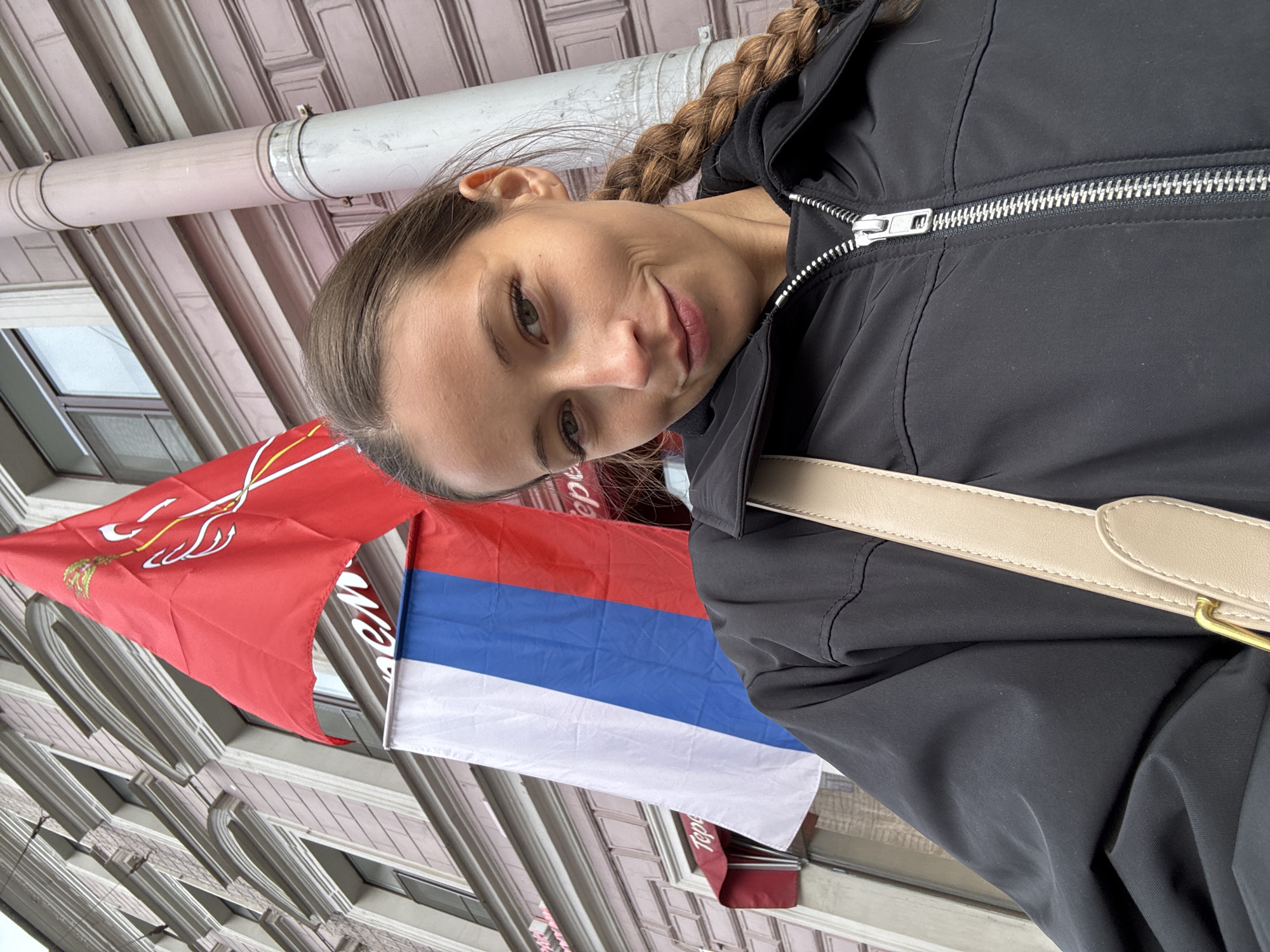 A woman with brown hair braided back is photographed close up, wearing a black jacket with a silver zipper and a beige purse strap visible over her right shoulder. In the background, a red flag with a white coat of arms and a Russian flag (white, blue, red stripes) hang from a building with a gray stone façade and arched windows. The photo appears to be taken in Saint Petersburg, Russia, as the red flag is the flag of the city of Saint Petersburg.