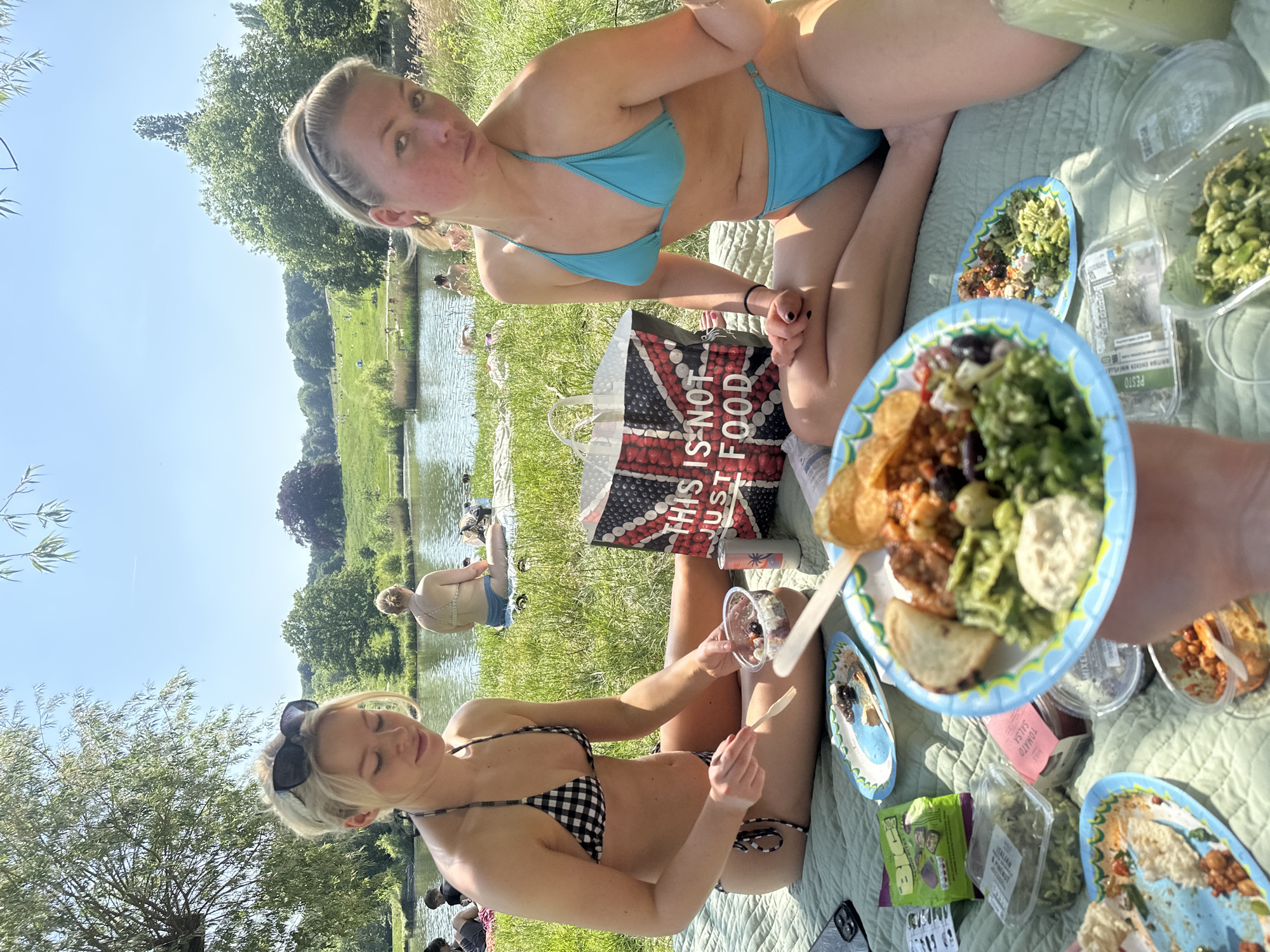 The image captures a picnic scene alongside a body of water, likely a pond or river, in what appears to be a park. Two blonde women, wearing bikinis, are sitting on a light green quilted blanket amidst long grass. One woman wears a black and white checkered bikini and sunglasses perched on her head, while the other is in a bright blue bikini. A shopping bag, branded "This is not just food" with the Union Jack design, is centrally positioned between them. Several paper plates and transparent food containers are scattered around, containing various foods. In the background, there's a grassy field with trees, and several individuals are either swimming or lounging by the water. The clear sky and bright sunlight suggest it's a summer day. Based on the image features and common knowledge, the location is likely Hampstead Heath, in London, United Kingdom.