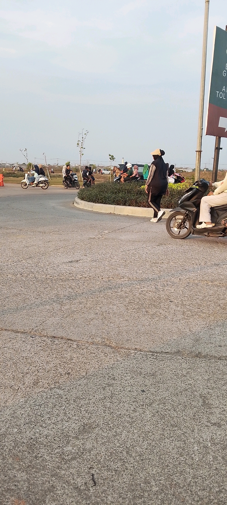In Satriamekar, Indonesia, an outdoor scene captures a group of people gathered near a landscaped area with low, dark green bushes. The ground in the foreground is a textured, light gray asphalt surface. Several motorcycles are present; one is being ridden by a person wearing white clothing and crocs, with its front wheel visible in the lower right corner. Another motorcycle, ridden by a person in black and white, is positioned to the left of the first, its front wheel turned towards the left. Further back, a group of individuals are seated or standing, some appearing to be on motorcycles. The sky above is a pale blue with a hazy, overcast appearance. A tall, white pole with a dark green and maroon sign is visible on the right side of the image. Trees with sparse foliage are scattered throughout the background.