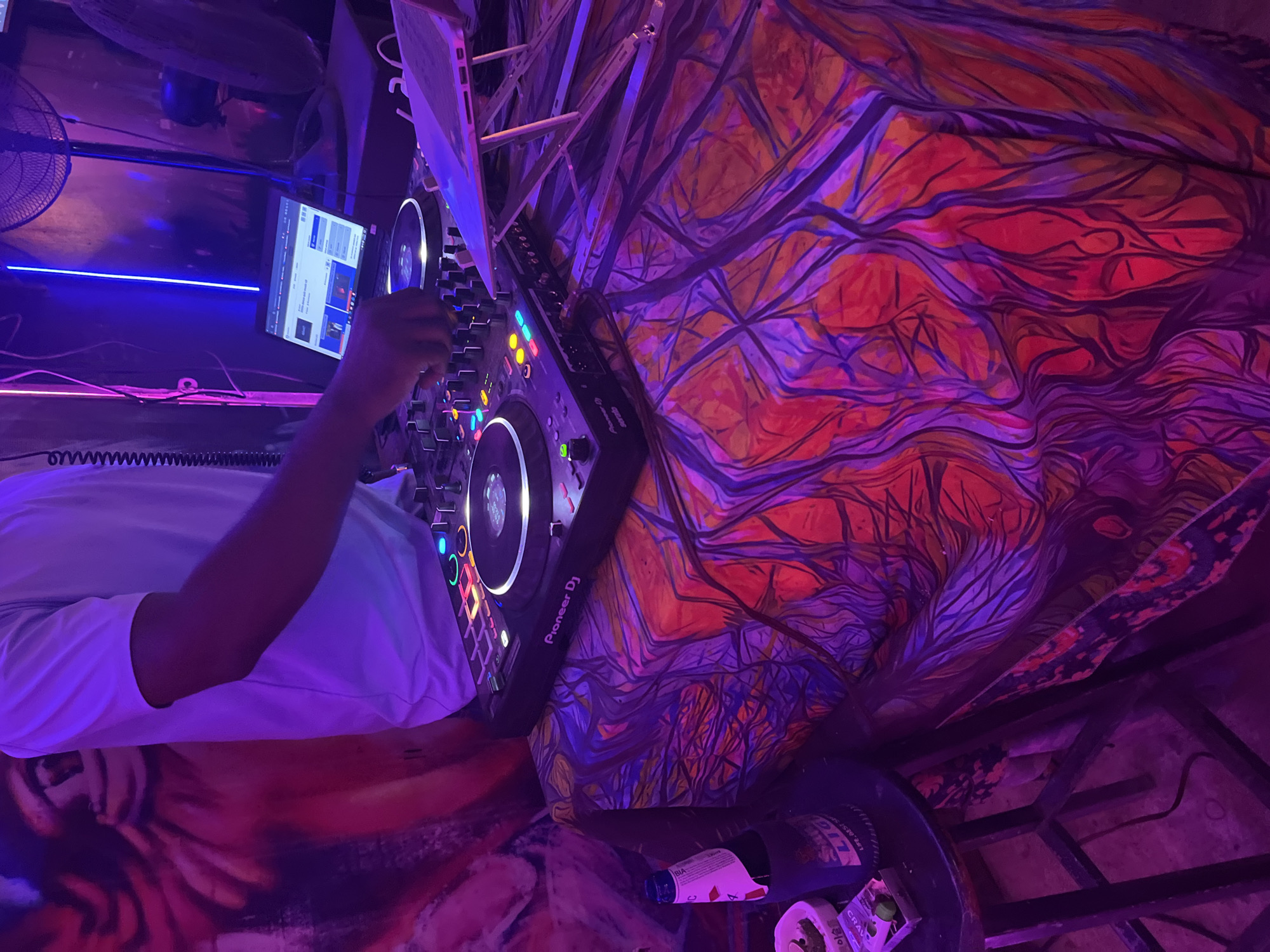 The image depicts a DJ operating equipment within a dimly lit venue in Đà Nẵng, Vietnam, bathed in purple, pink, and blue neon lighting. A person wearing a white short-sleeved shirt is visible from the torso up, with their right hand adjusting a knob on a black Pioneer DJ Serato controller. The controller features illuminated pads and buttons in various colors including green, blue, red, orange, and yellow. To the right of the controller, a silver laptop on a stand displays a web browser. The DJ equipment rests on a table covered with a vividly patterned cloth in shades of purple, red, and orange, resembling abstract organic forms. In the lower left foreground, a dark circular stool holds a bottle of "Light Beer," a white ashtray containing cigarette butts and a lime wedge, and a small package labeled "CRAV." The background on the left features a large reddish-purple wall mural depicting an abstract, stylized face. On the right, vertical blue and pink neon strip lights are visible, with dark industrial-style fans positioned behind them.