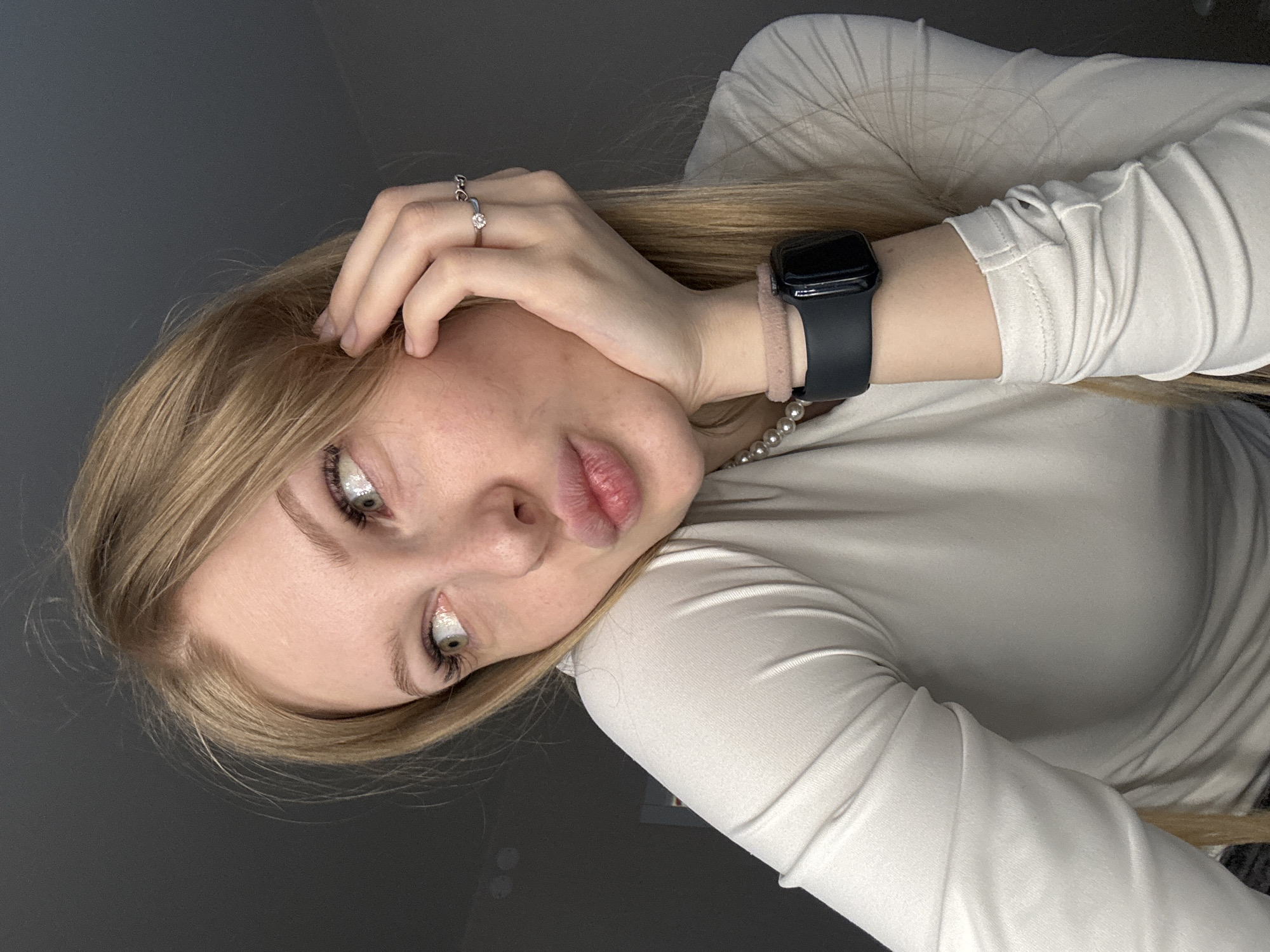 A portrait-style photograph, originally rotated 90 degrees clockwise, features a young woman with long blonde hair and fair skin. She is positioned looking directly at the camera, with her head slightly tilted and lips gently puckered in a playful expression. Her left hand is raised to her head, with fingers lightly touching her hair. On her left hand, two silver-colored rings are visible: one with a small clear stone on her ring finger, and a plain band on her middle finger.
Her left wrist displays a black smartwatch with a matching black band, and a light pink or nude-colored scrunchie is worn beneath it. A string of pearls from a necklace is partially visible around her neck. Her eyes are a prominent feature, enhanced with glittery or iridescent makeup on the inner corners and lower lash line, along with long, dark eyelashes.
She is wearing a light-colored, long-sleeved top or sweater. The background is a plain, dark gray surface, indicating an indoor setting. The lighting is soft and appears to be frontal, illuminating her face and upper body. The image captures a casual, posed moment. No text is visible within the photograph. The context for this image is Perm, Russia.