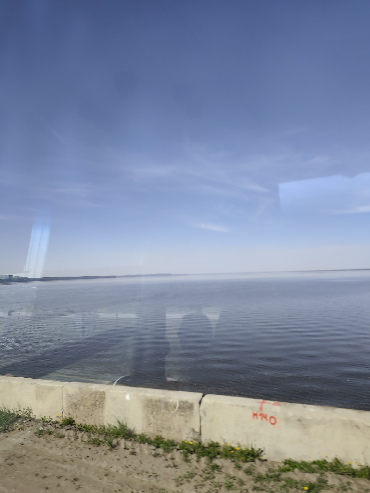 The image depicts a body of water, likely a river or lake, under a blue sky with thin clouds. A concrete barrier is in the foreground, with grass and small yellow flowers growing near it. Red graffiti "M40" is visible on the barrier. The water has gentle ripples, suggesting a slight breeze. The scene is a view from a car, possibly along a road or highway bordering the water. Given the location of Chaikovsky, Russia, it is possible that the body of water is the Votkinsk Reservoir. The time of day is likely daytime, indicated by the bright sky.
