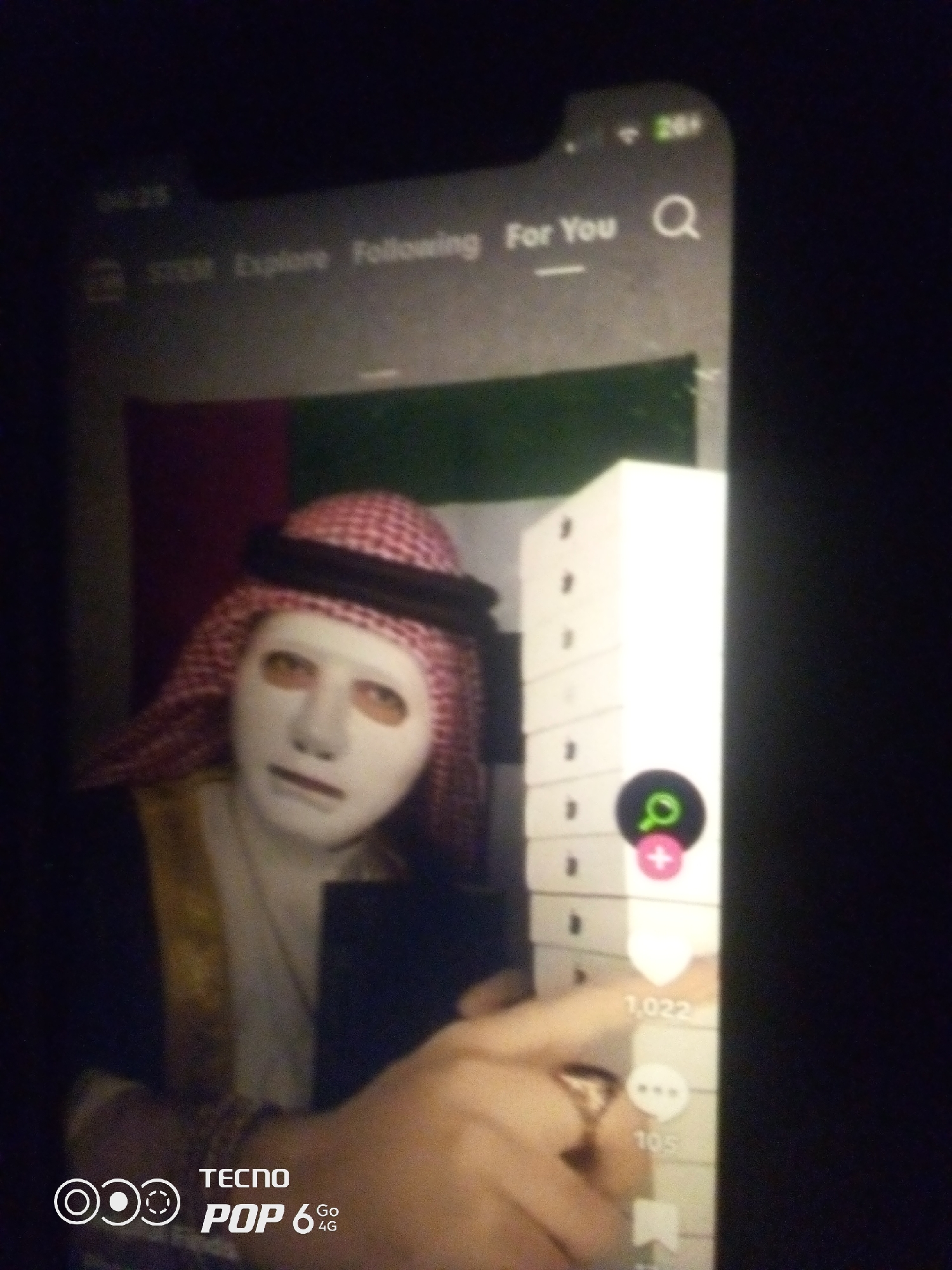 A person wearing a white mask and a red and white checkered keffiyeh is holding a white smartphone displaying a TikTok video. The video shows a person in traditional Arabic clothing, with a white mask covering their face and orange-tinted eyeholes. To the right of the masked person in the video, a tall stack of white boxes is visible, with social media icons and engagement numbers (1,022 likes and 105 comments) displayed on the side. The smartphone screen also shows the TikTok interface with options like "Following" and "For You." In the bottom left corner of the smartphone screen, the TECNO POP 6 Go 4G logo is present. The overall image is dark, suggesting it was taken in low-light conditions. There is no indication of the city of Howayi, Nigeria.