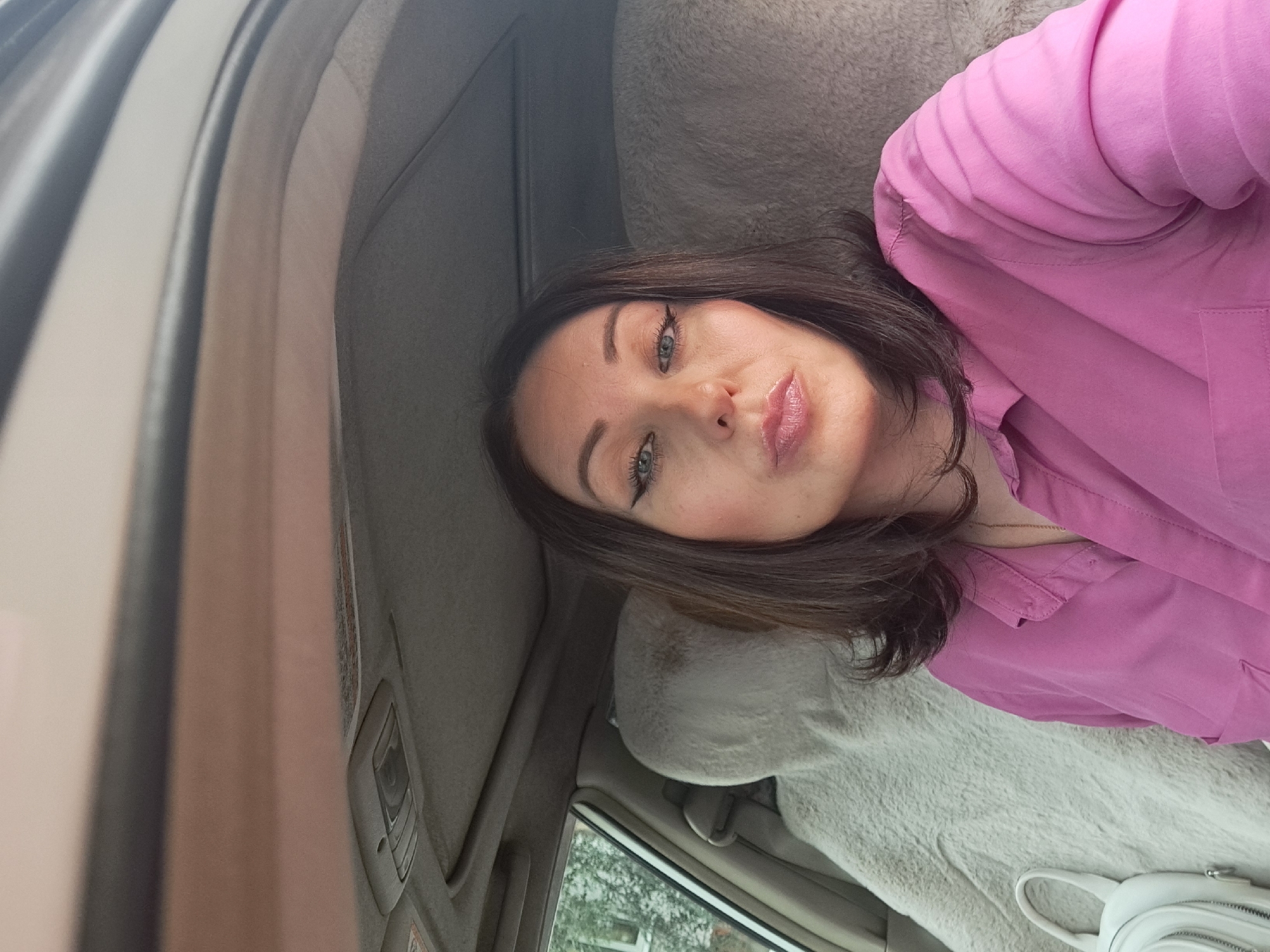 A woman is sitting in the passenger seat of a car, looking at the camera and puckering her lips. She has dark brown hair and is wearing a pink collared shirt. A fluffy grey seat cover is visible behind her. The interior of the car, including the ceiling and a small bag on the seat, is also visible. The scene appears to be taking place during the daytime.
