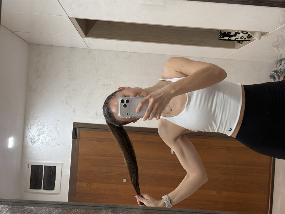 A female subject is depicted in a mirror selfie taken indoors. She wears a white athletic crop top, black leggings, a silver necklace with a small pendant, and a white smartwatch on her left wrist. Her dark brown hair is styled in a high ponytail, which she holds with her left hand, simultaneously flexing her left bicep. Her face is largely obscured by a silver-colored smartphone, held in her right hand, used to capture the image. A subtle smile is visible on the lower right portion of her face.
The immediate background, reflected in the mirror, features a dark brown wooden door with a metallic security plate to the left of the subject. Above the door, on a textured, light-colored wall, a white electrical panel with two horizontal dark circuit breakers is mounted. To the subject's right, white, glossy, floor-to-ceiling wardrobe doors are visible. A recessed open shelf within the cabinetry contains a folded item with a black and white leopard print pattern. Grey and white athletic shoes are partially visible on the floor in the bottom right corner. The environment appears to be an interior space, likely a private residence. The specific location within Varna, Bulgaria, is not discernible from the visual evidence.
