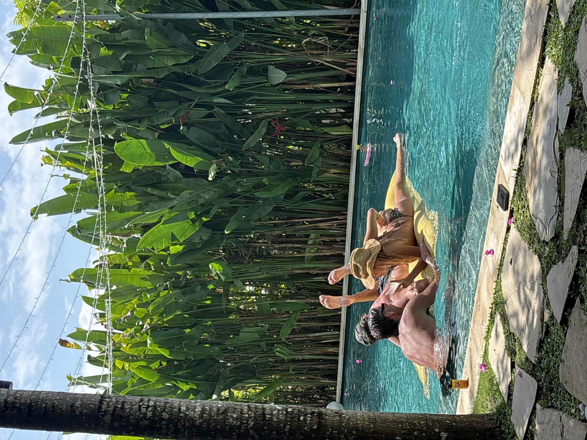 A horizontal eye-level shot shows a couple in a pool at Kuta Selatan, Indonesia. A woman wearing a bikini and a hat lies on a yellow inflatable float in the pool, while a man is in the water with her. The pool is bordered by greenery and a stone-tiled area. Tall plants with large leaves are visible in the background, with string lights hanging above them. A palm tree trunk is in the foreground. The sky is partially visible with clouds.