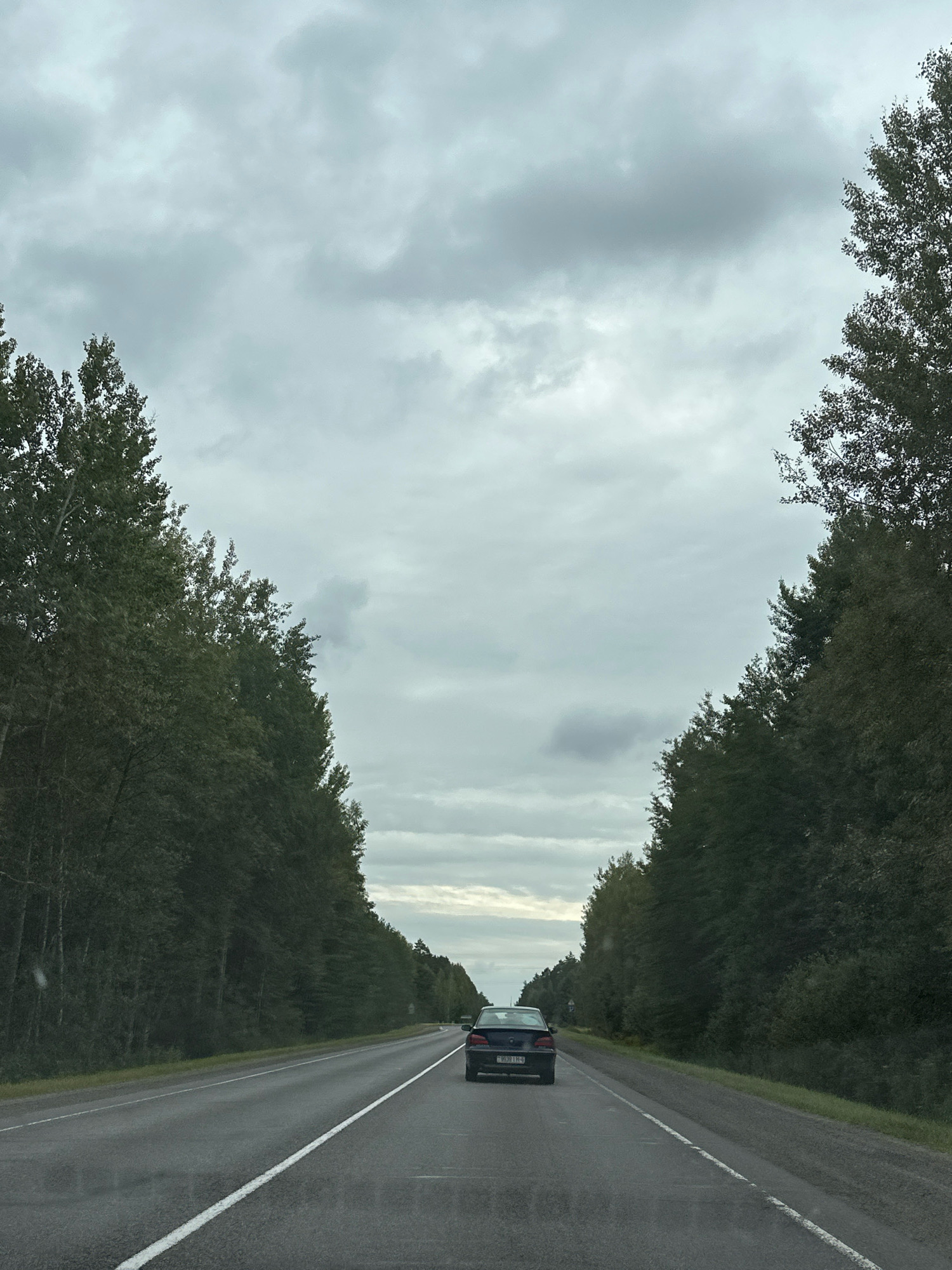 The image depicts a grey, overcast sky above a two-lane asphalt road. Tall, dense trees line both sides of the road, creating a tunnel-like effect. A dark-colored sedan is driving away from the viewer down the right lane, its license plate visible. The road surface appears wet, suggesting recent rain. The overall scene is one of driving through a wooded area under a cloudy sky. The specific location within Гарбацэвіцкі сельскі Савет, Belarus cannot be determined from the visual information alone.