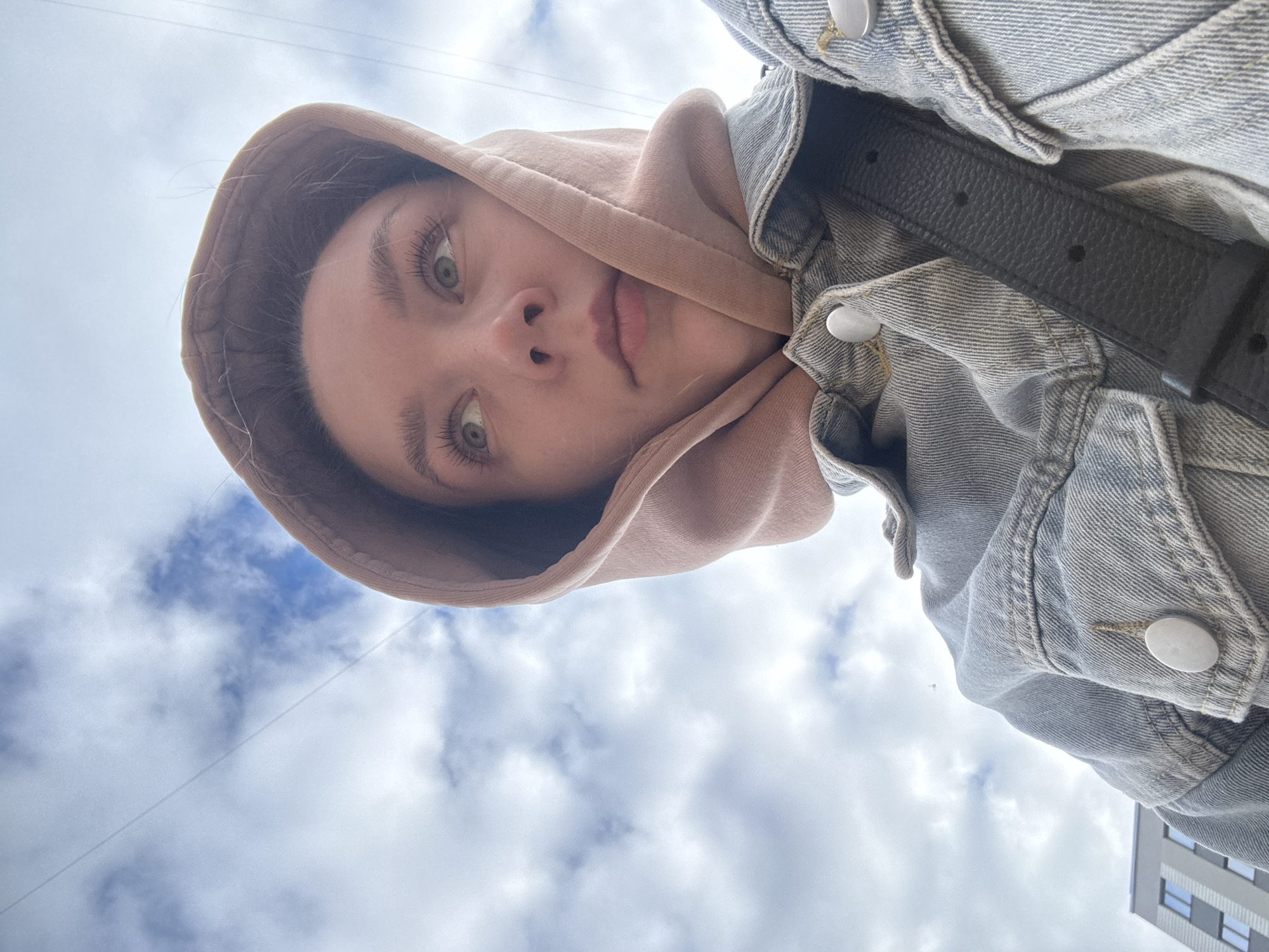 A low-angle, upward-facing portrait taken in Izhevsk, Russia, features a young individual with fair skin, light green eyes, and light brown hair partially concealed by a light brown hooded sweatshirt. The individual is wearing a light blue denim jacket with prominent silver buttons and a black textured leather strap across the upper chest, visible over the jacket. The subject looks directly into the camera. The background consists primarily of an overcast sky with white and grey clouds and patches of blue. In the upper left, a segment of a modern multi-story building with a light-colored facade and dark rectangular windows is visible. Several thin, dark linear structures, likely overhead wires, traverse the sky in the upper right quadrant of the image.