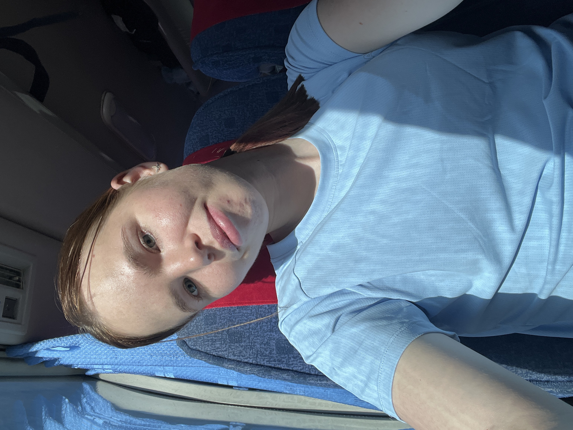 A fair-skinned woman with auburn hair in a ponytail is in an automobile. The woman is wearing a blue t-shirt. She is sitting on a red and blue patterned car seat with blue fabric below. The car's interior and exterior are visible, with a car door panel in the lower left. The image does not contain sufficient information to identify the specific location within the city of Berezniki, Russia.