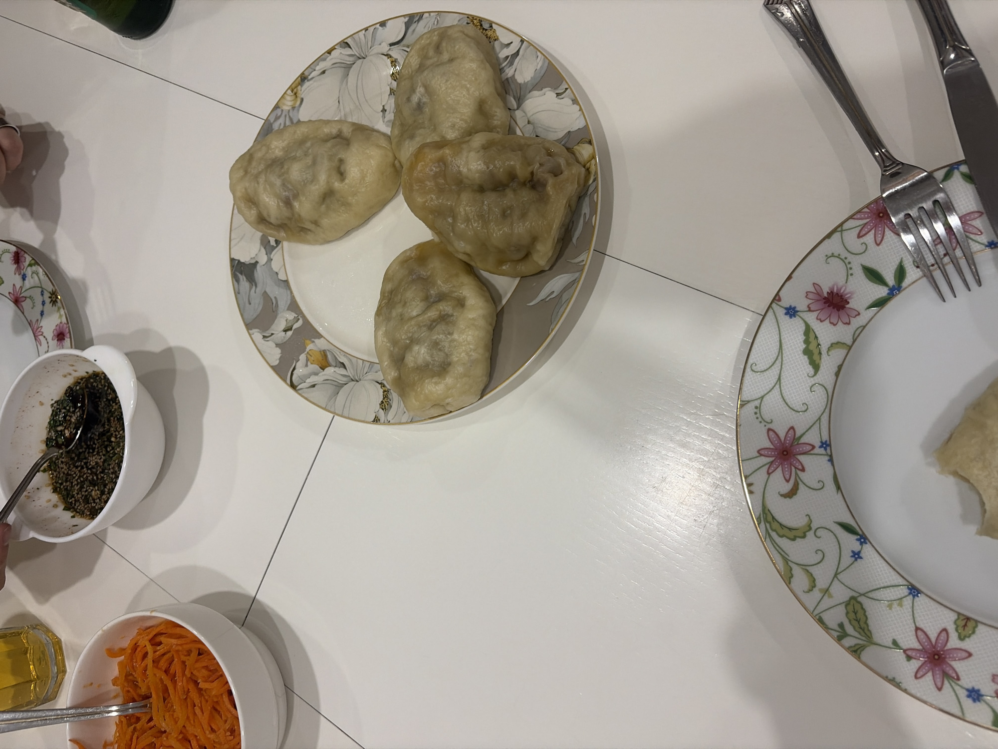 A top-down shot shows a table set with food and utensils. A floral-rimmed plate holds four dumplings. Nearby, a bowl contains a dark, sesame-based sauce with a spoon inside, and another bowl holds shredded orange carrots with chopsticks resting on top. To the right, another floral-rimmed plate has a knife and fork placed on it. The table is white, suggesting a clean and modern environment. Without any other information, it's impossible to determine if the location is in Saint Petersburg, Russia, or any other city for that matter.
