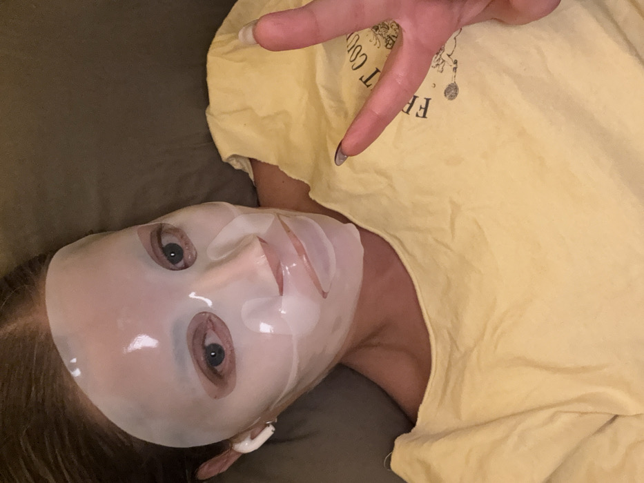 A woman wearing a translucent sheet face mask is lying on a soft, gray surface. She's angled towards the camera, wearing a yellow t-shirt with a printed design. One hand is raised, fingers forming a peace sign. An earbud is visible in her left ear. The mask covers her face entirely, with openings for the eyes, and is shaped to the contours of her nose and mouth.