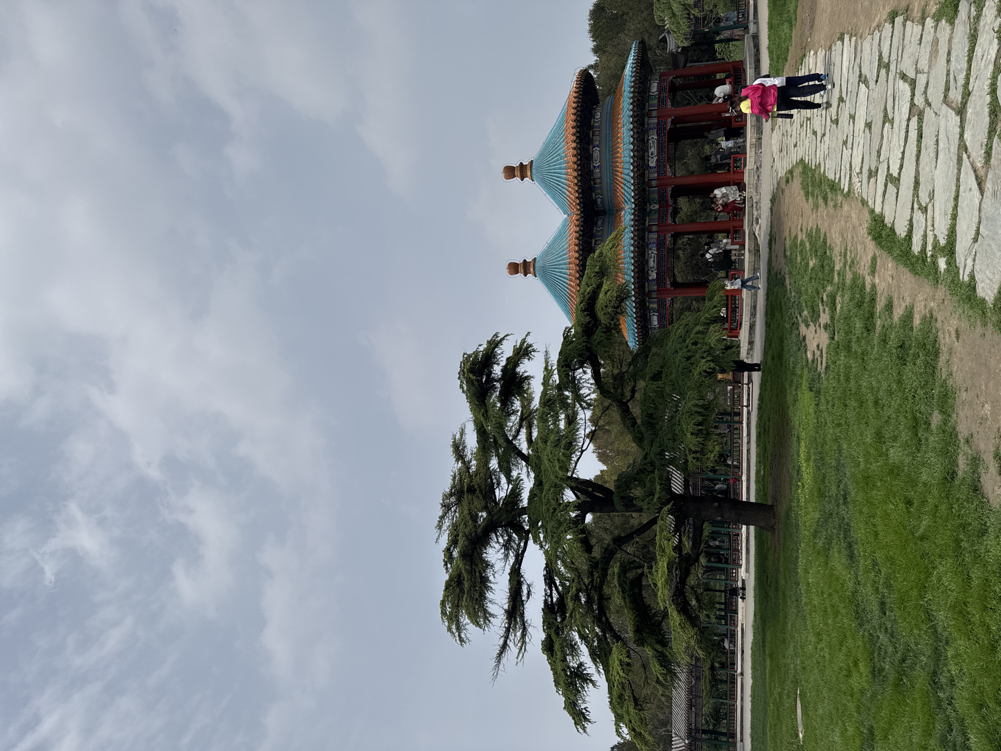 The image depicts a scene in what appears to be a park in Dongcheng District, China. In the foreground, a large, mature tree with dense foliage dominates the view. To the right of the tree, a traditional Chinese structure with a multi-tiered roof in blue and orange hues is visible. People are present within and alongside the structure. A pathway paved with stone slabs runs along the right side of the frame, with two individuals walking on it. The environment consists of a green lawn. Above, the sky is overcast with a light, cloudy cover.