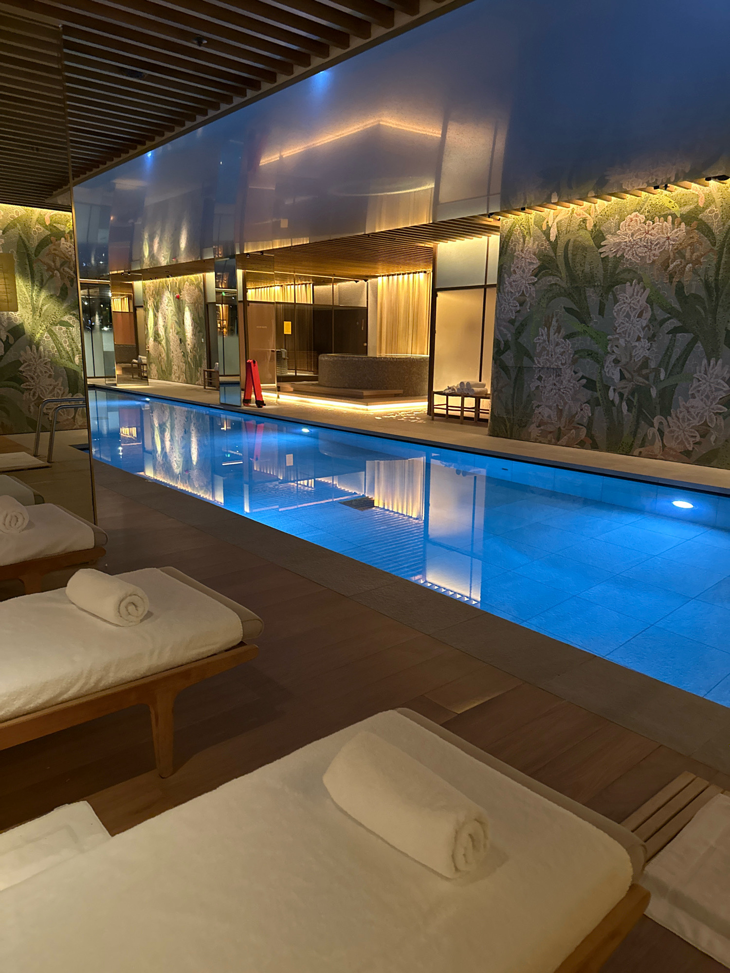The image shows an indoor swimming pool area, likely within a luxury hotel or spa, in London, United Kingdom. The pool is illuminated with blue underwater lights, casting reflections on the ceiling and walls. Along the edge of the pool are several lounge chairs, each with a white rolled towel neatly placed on it. The walls are decorated with a floral mosaic pattern and large mirrors, creating a sense of spaciousness and elegance. The ceiling features a modern design with wooden slats and soft, ambient lighting. In the background, there is a reception or changing area with curtains and further lighting. There are no people visible in the image.