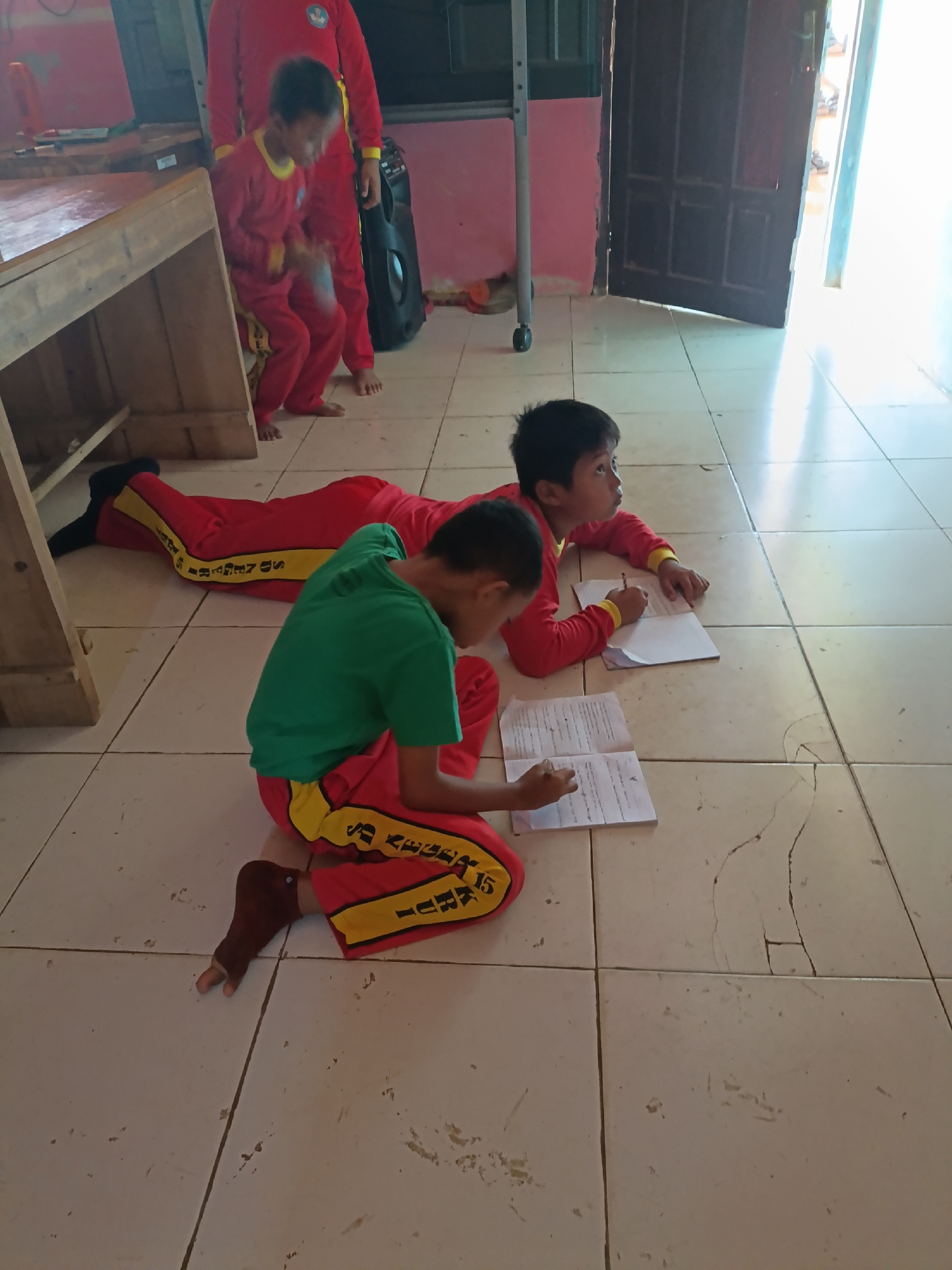 In a room with a tiled floor, possibly in the city of Sumberejo, Indonesia, two young boys are engaged in writing on separate notebooks. One boy, wearing a green shirt and red pants with yellow trim, is positioned closer to the foreground, kneeling on the floor and intently focused on his notebook. His right foot is visible, with a dark brown covering that appears to be a sock or a protective wrap. The other boy, lying on his stomach and wearing red attire with yellow trim, is further back in the frame. He is also writing in his notebook, with his head turned slightly to his left. His arm is extended, and he holds a pencil. In the background, another child or adult, also dressed in red, stands partially visible, their lower body in view. A black speaker unit is positioned behind the standing figure. A wooden table with shelving is present to the left of the frame, and a doorway with a dark wooden door is visible in the background to the right. The lighting suggests it is daytime, with sunlight streaming in from the right side of the room. The overall scene depicts a study or classroom environment.
