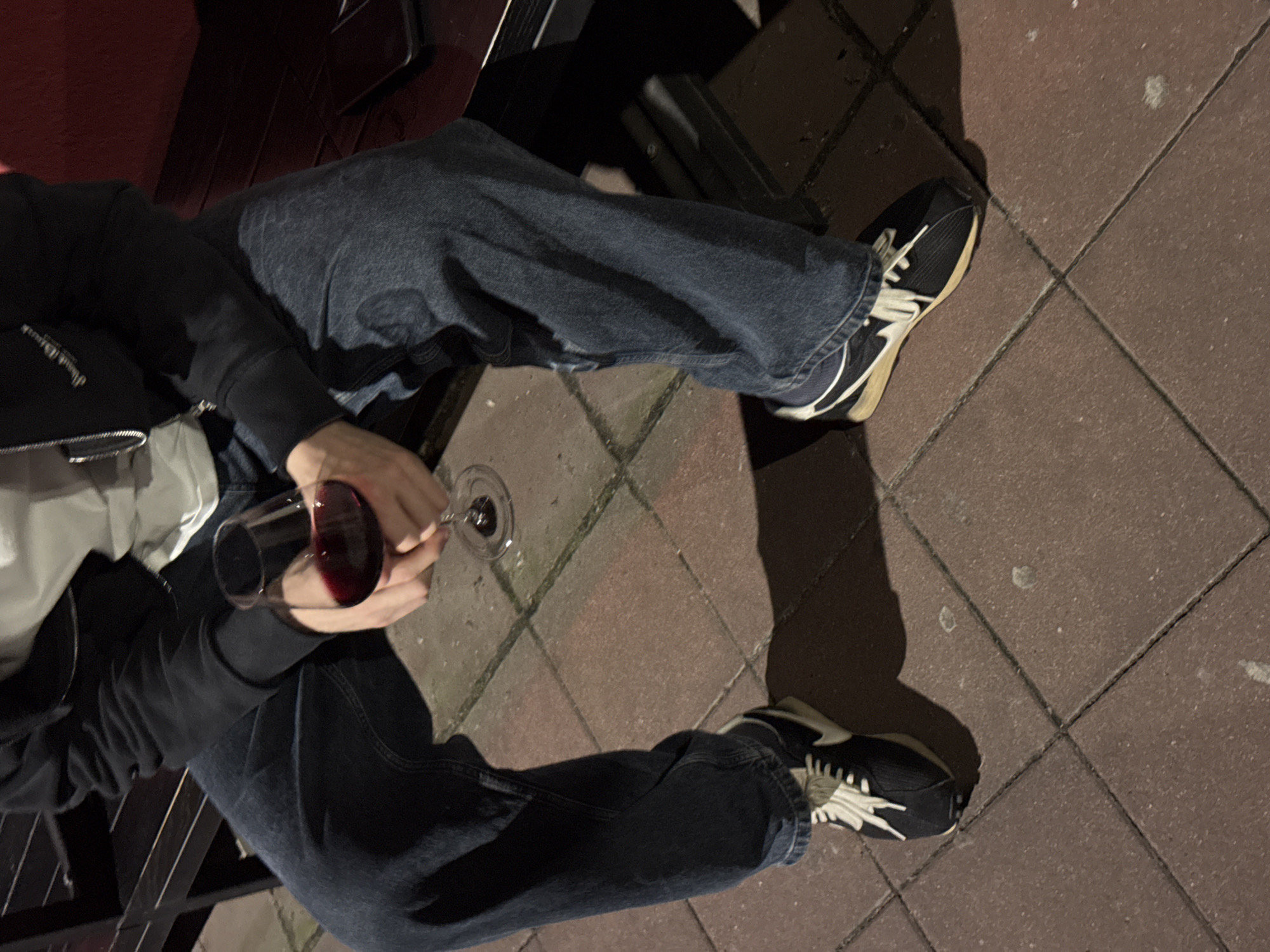 A person sits on a park bench, dressed in blue jeans, black and white sneakers, a white t-shirt, and a black hoodie. They are holding a glass of red wine. The setting appears to be outdoors on a paved surface, possibly a public area in Belgrade, Serbia. The time of day appears to be evening due to the lighting and shadows.